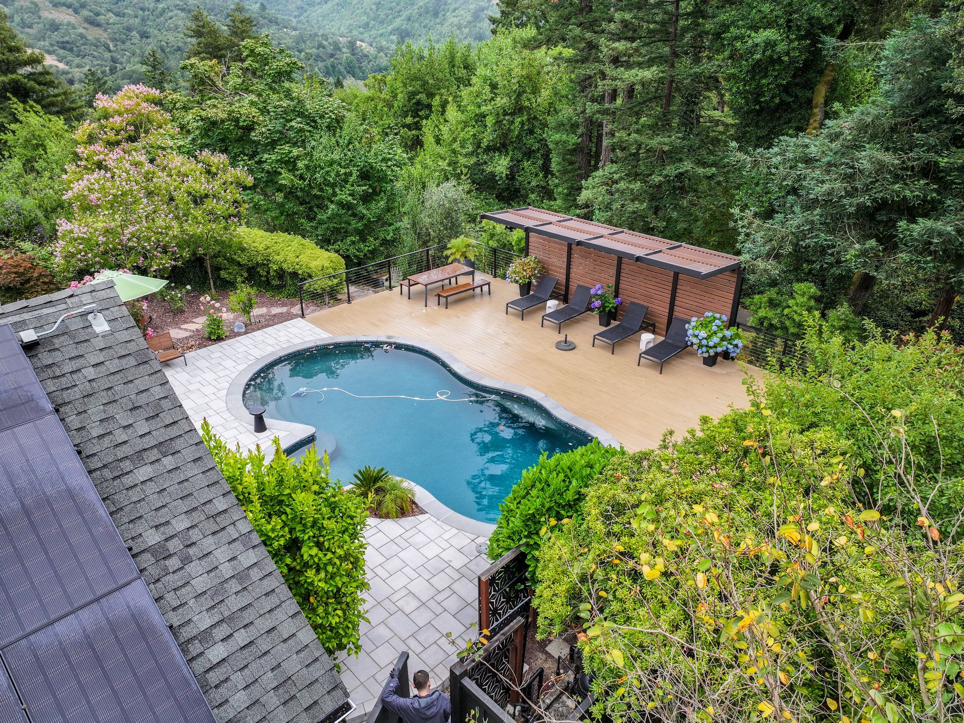 Backyard pool with tan patio, pergola seating, and lush green hillside trees