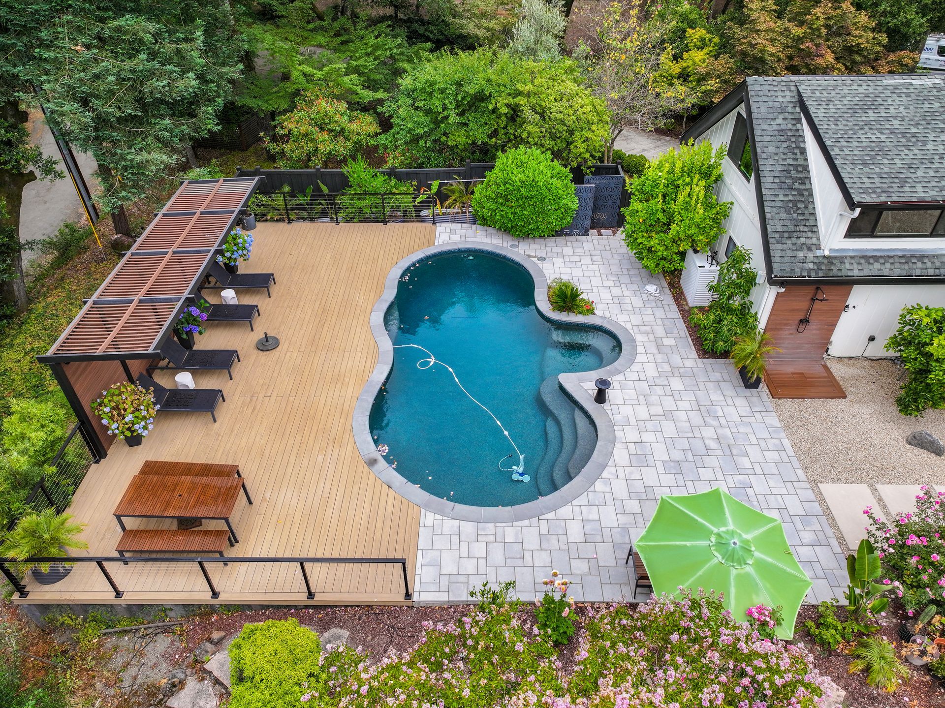 Aerial view of a backyard pool with lounge chairs, pergola seating, and an umbrella surrounded by greenery