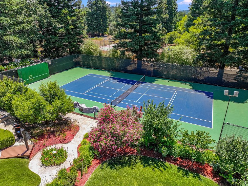 Outdoor tennis court with blue surface, green fencing, and landscaped garden surrounding it