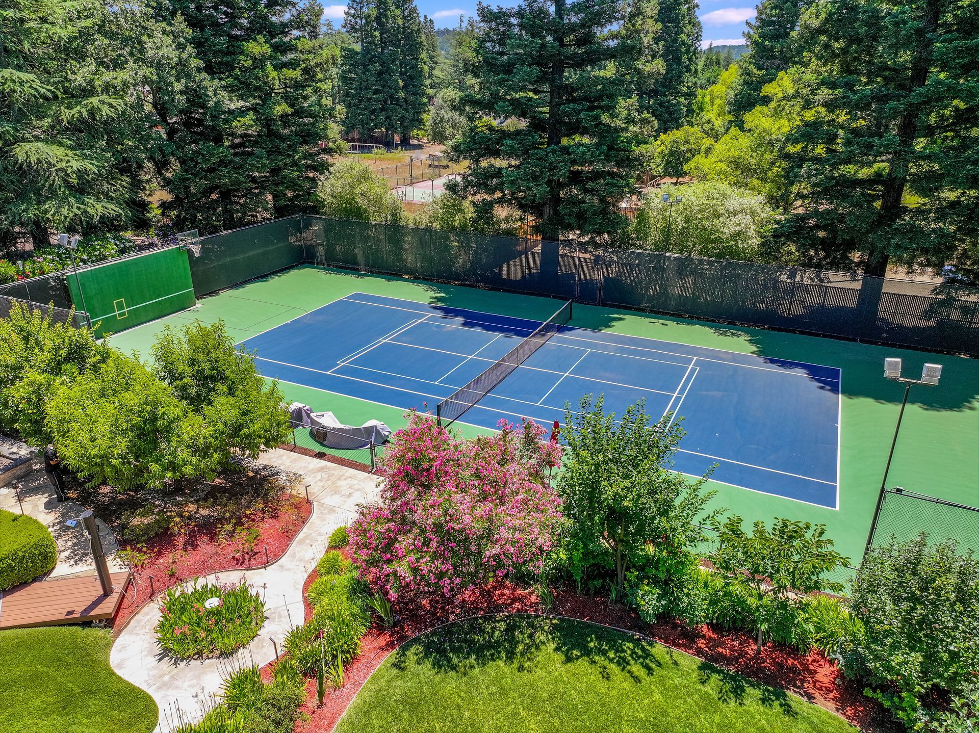 Blue tennis court surrounded by green fences, lush trees, and landscaped garden beds under bright sunlight