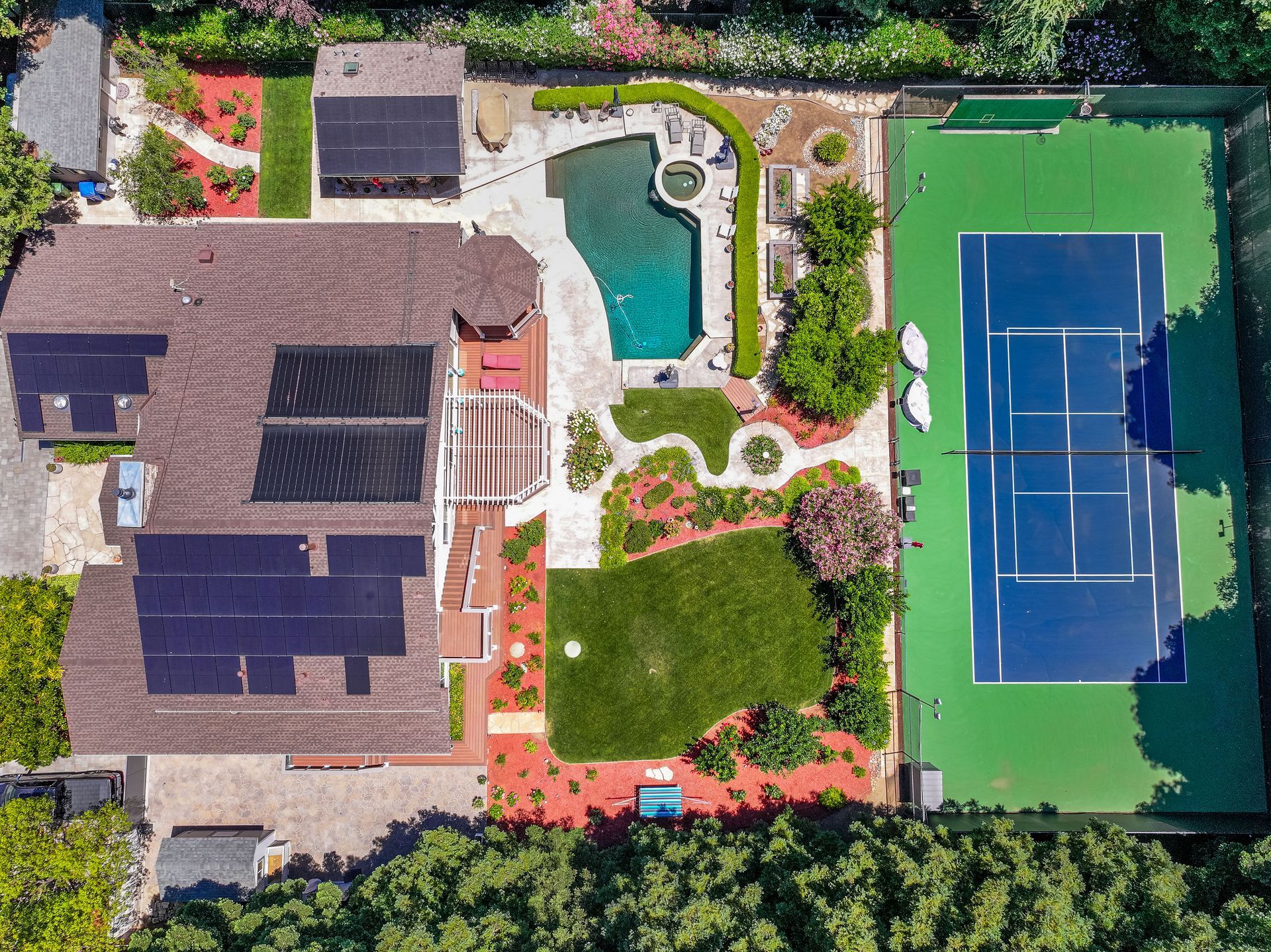 Aerial view of a house, pool, garden, and tennis court in a landscaped backyard