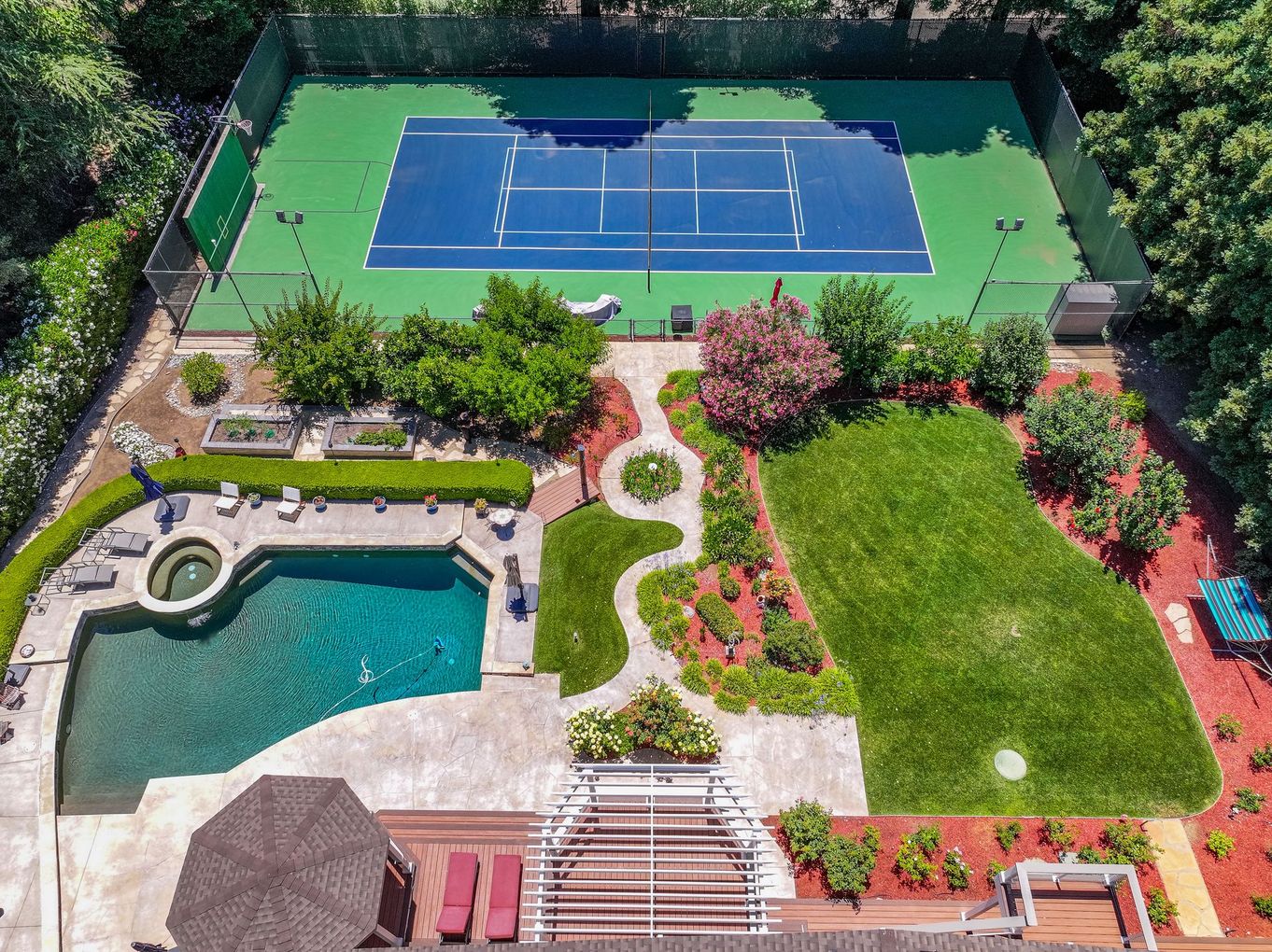 Aerial view of a backyard with a pool, patio, garden, and a blue tennis court.