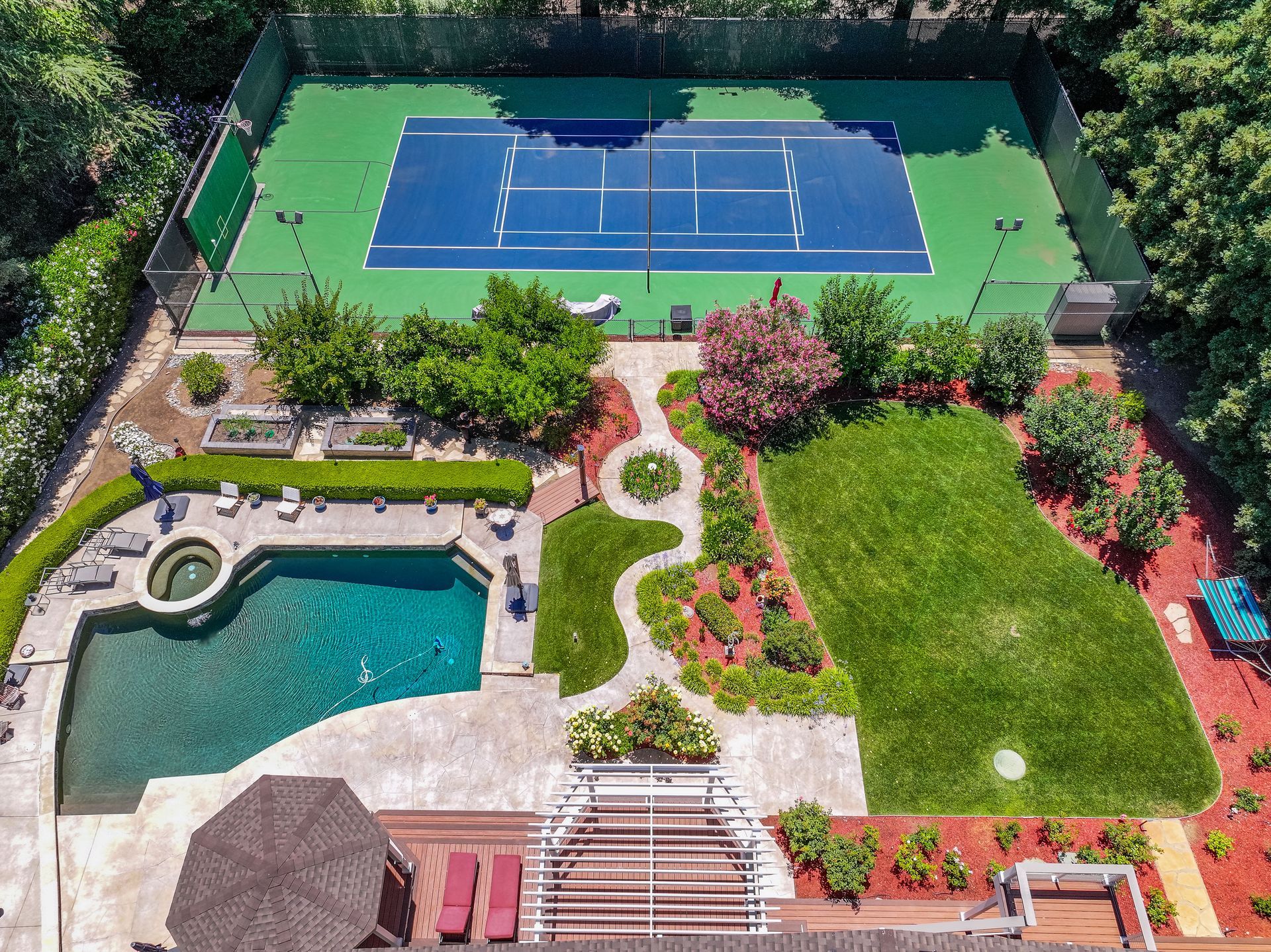 Aerial view of a backyard with a swimming pool, patio, lawn, and tennis court surrounded by trees.
