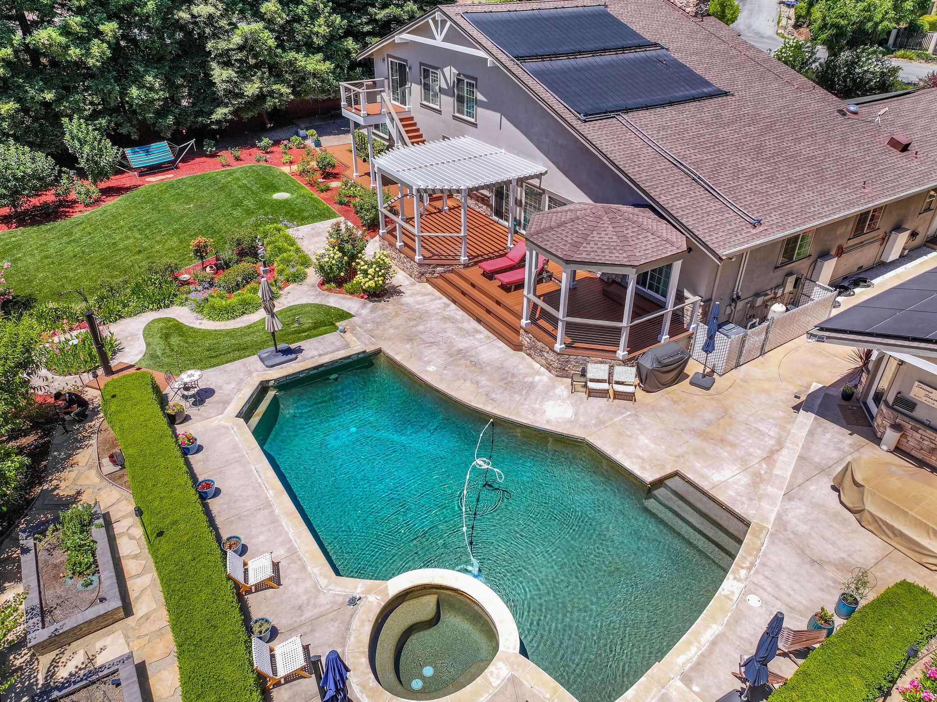 Aerial view of a backyard pool, hot tub, patio, and house with landscaped garden.