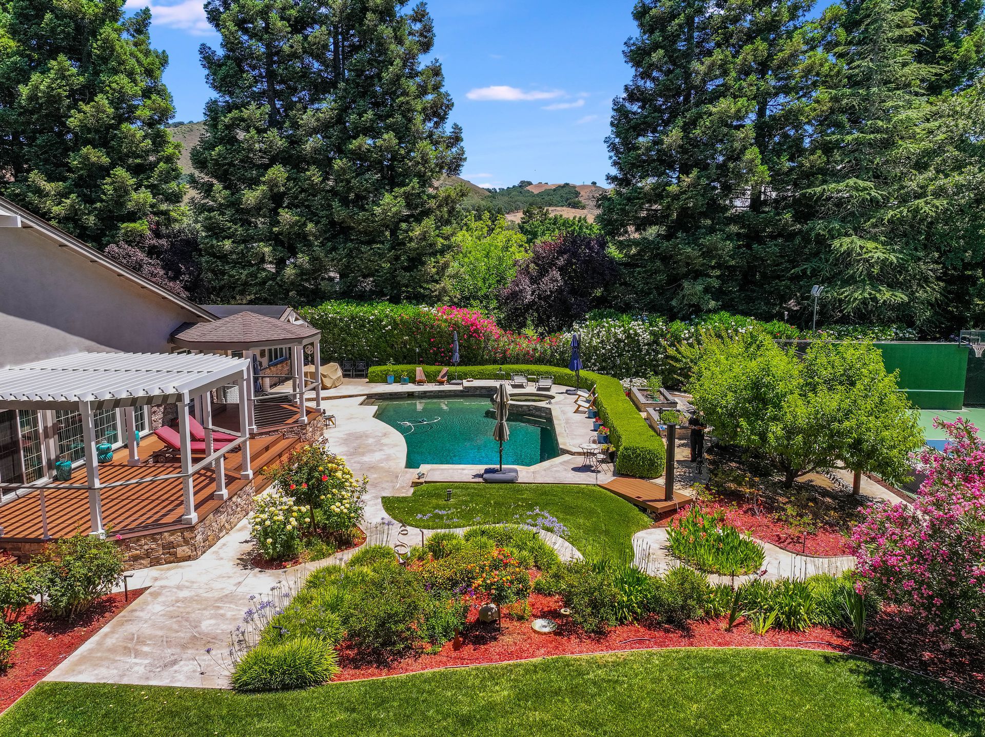 Backyard with a swimming pool, patio, and colorful flower beds surrounded by tall trees