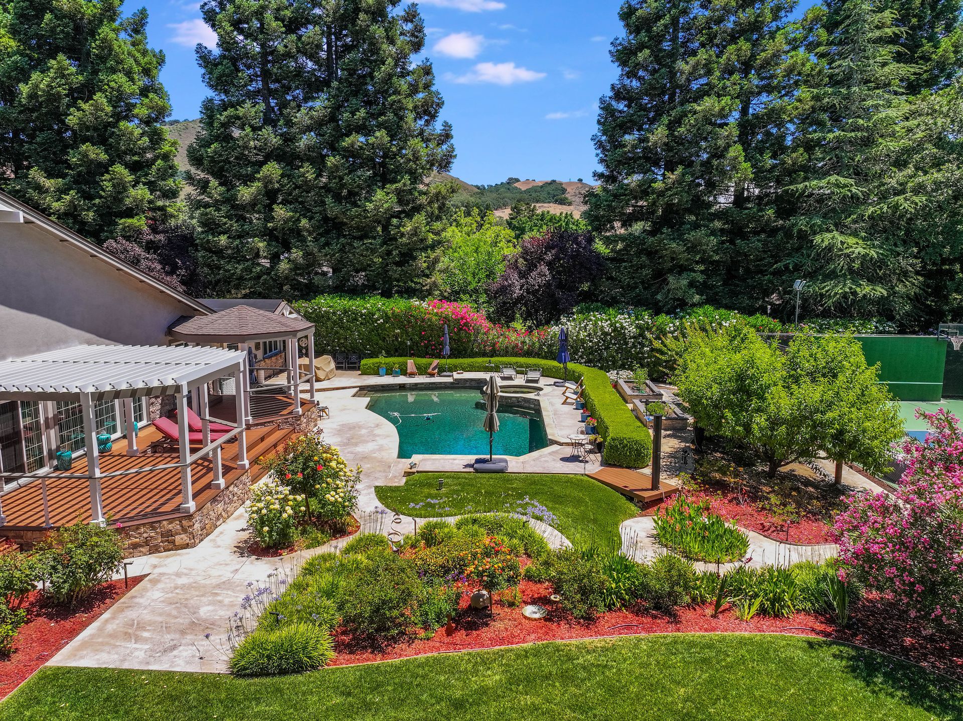 Backyard with pool, patio gazebo, and colorful flower beds surrounded by tall trees