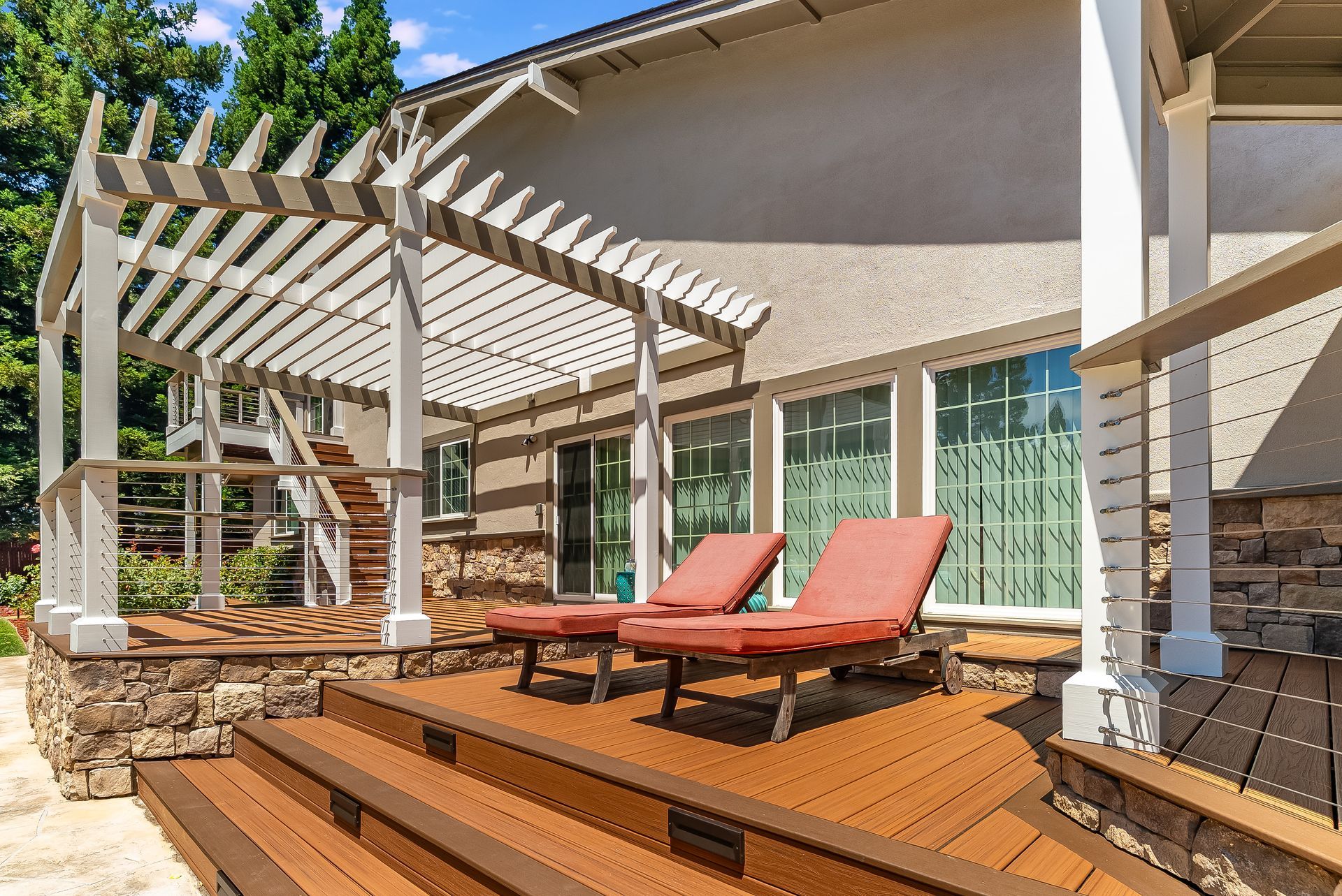 Backyard deck with pergola, red lounge chairs, and sliding glass doors beside a stone planter