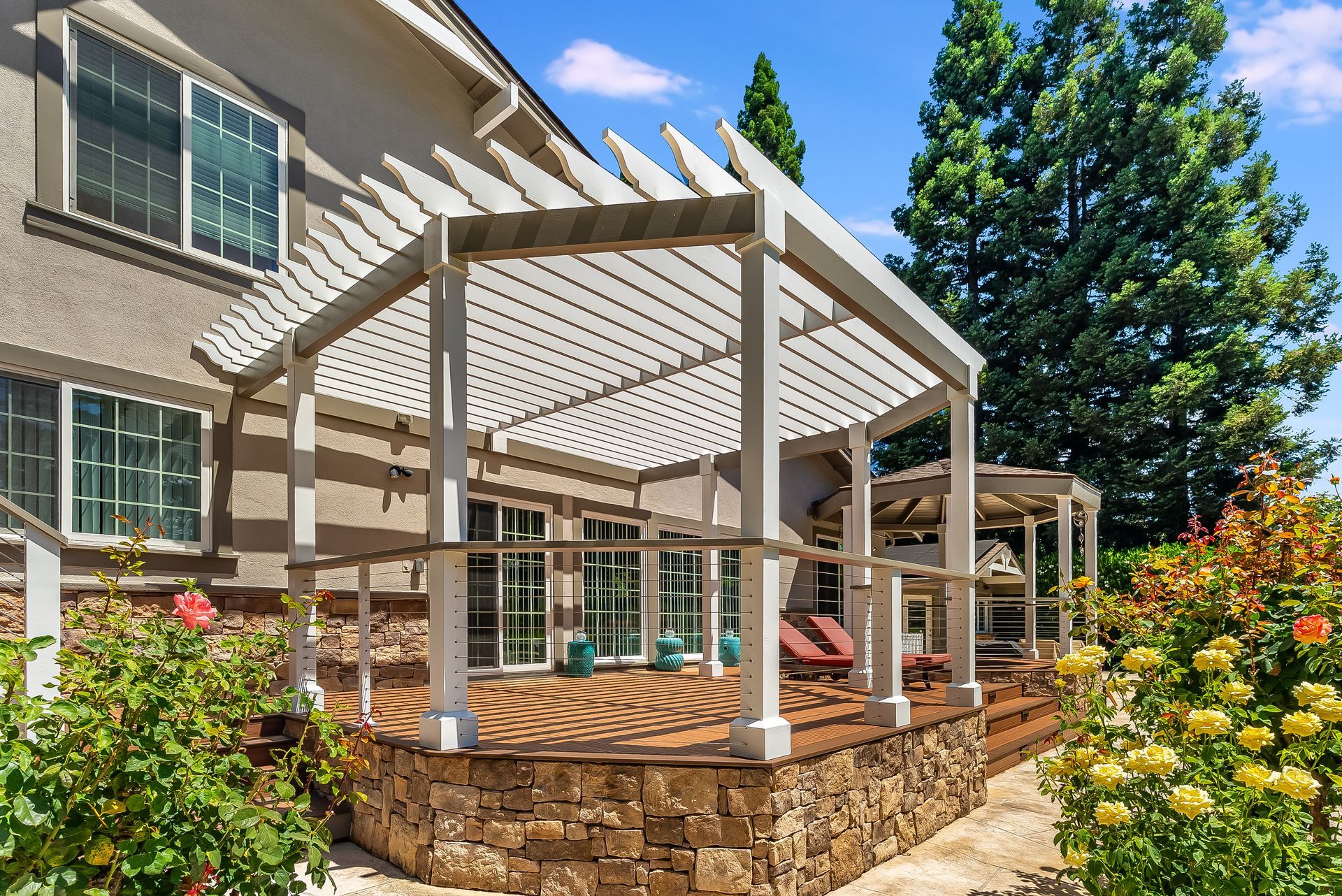 Stone patio with white pergola beside a brick house and garden flowers