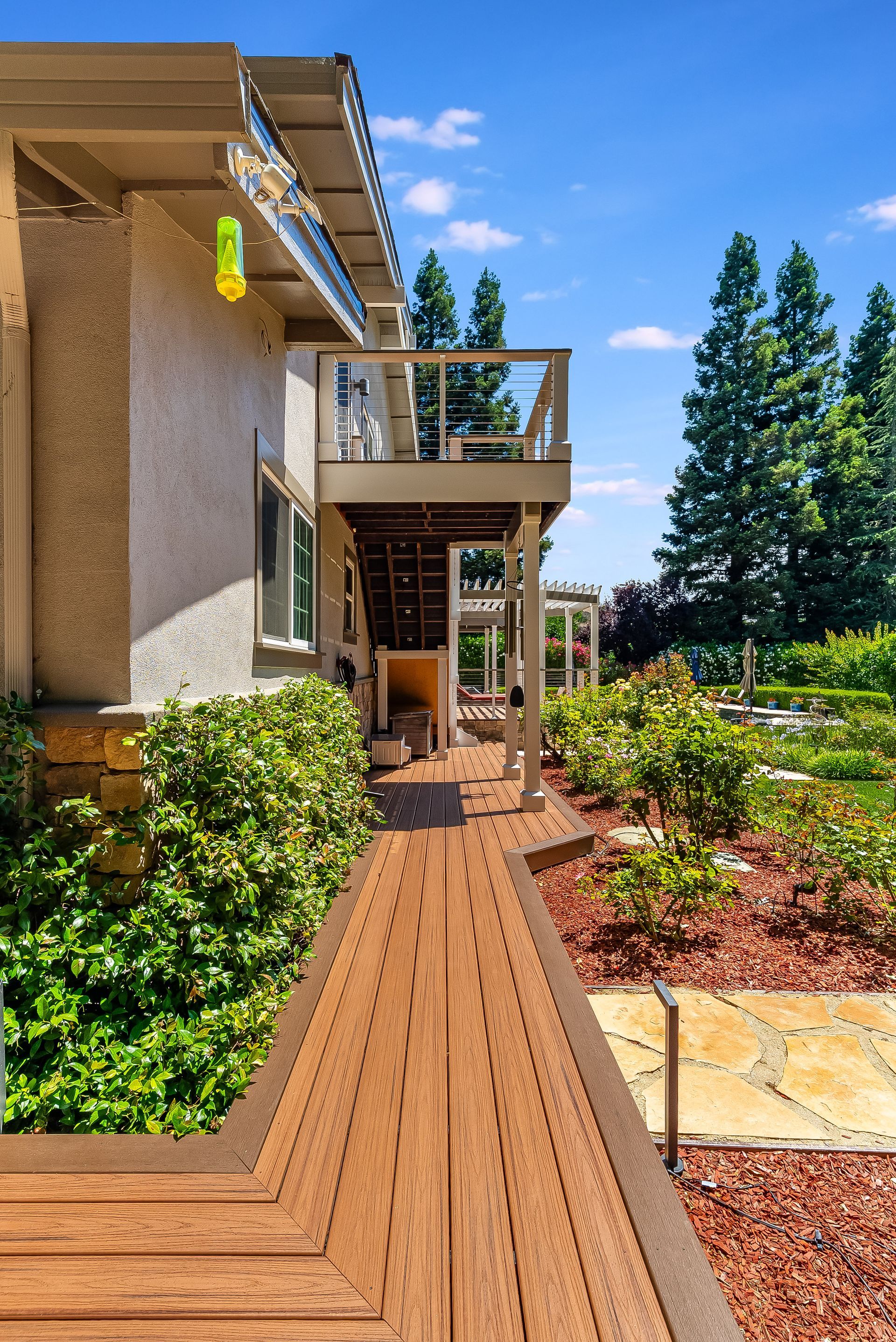 Sunlit garden walkway beside a beige house with wooden deck and green shrubs
