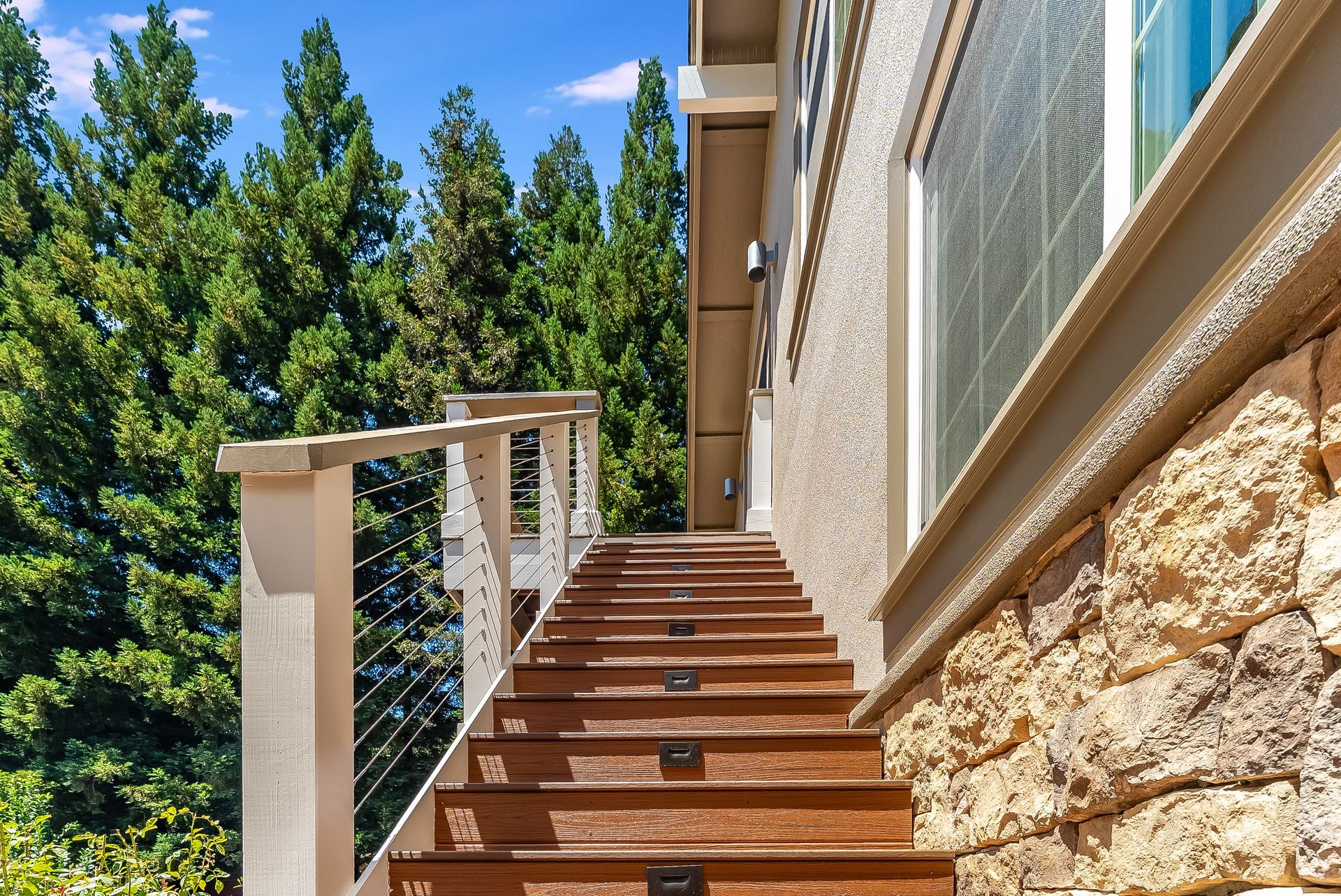 Outdoor wooden staircase beside a stone house, with white railings and green trees in the background