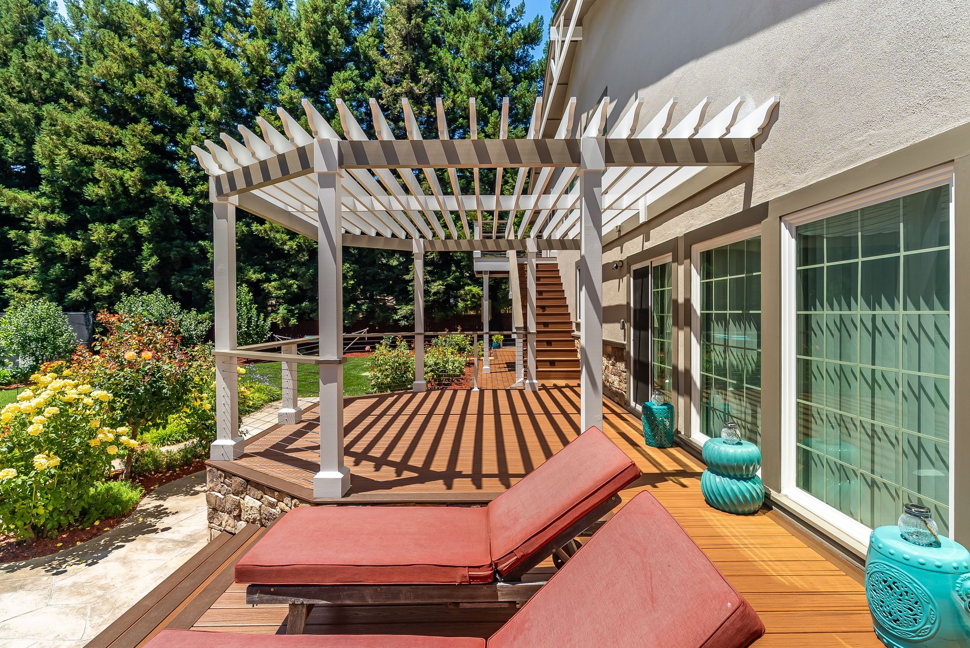 Backyard patio with white pergola, wooden deck, red cushions, potted plants, and sliding glass doors