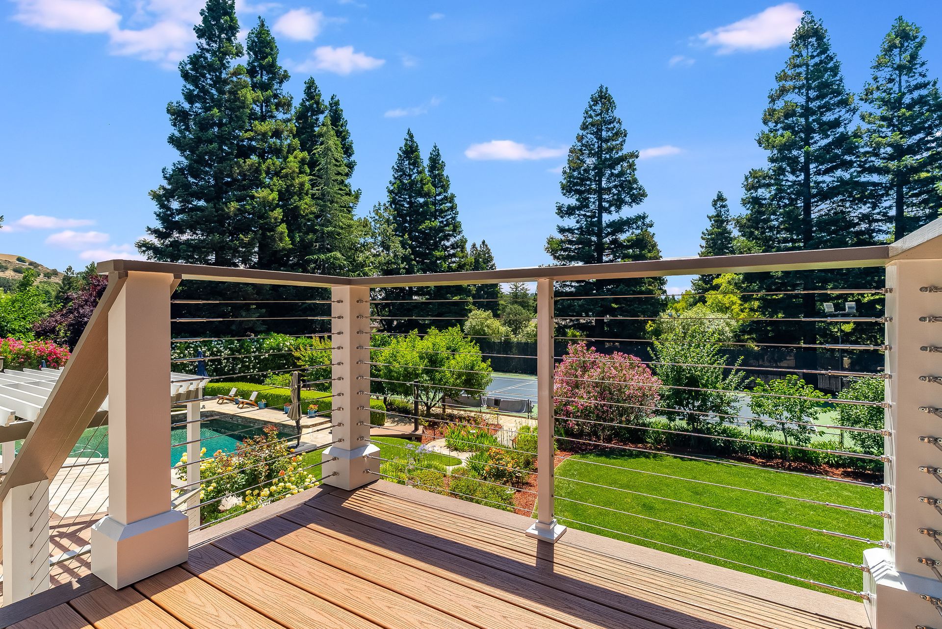 Wooden deck overlooking a landscaped garden with green lawn, shrubs, and tall pine trees under a blue sky
