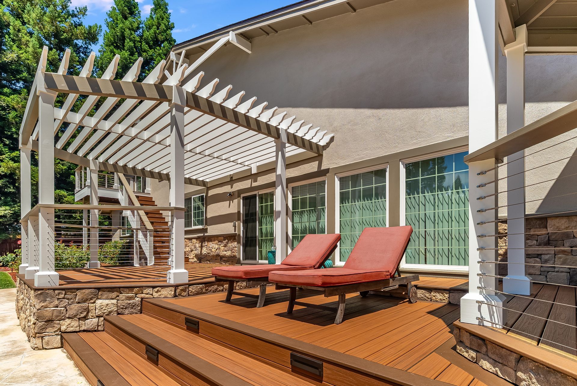 Wooden backyard deck with red lounge chairs and a white pergola beside sliding glass doors