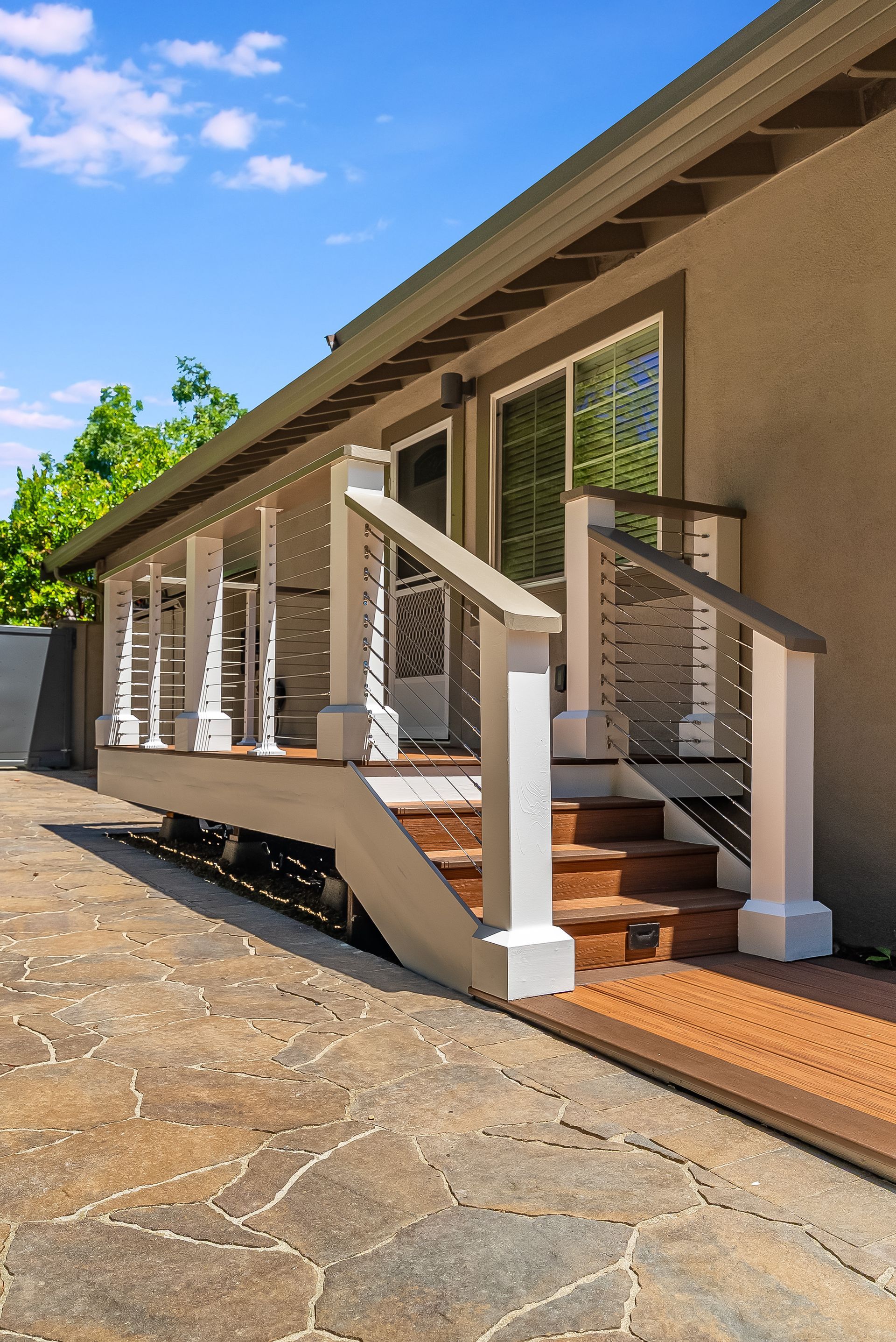 Sunlit exterior of a tan house with a long front porch, white railings, and brick steps on a stone walkway