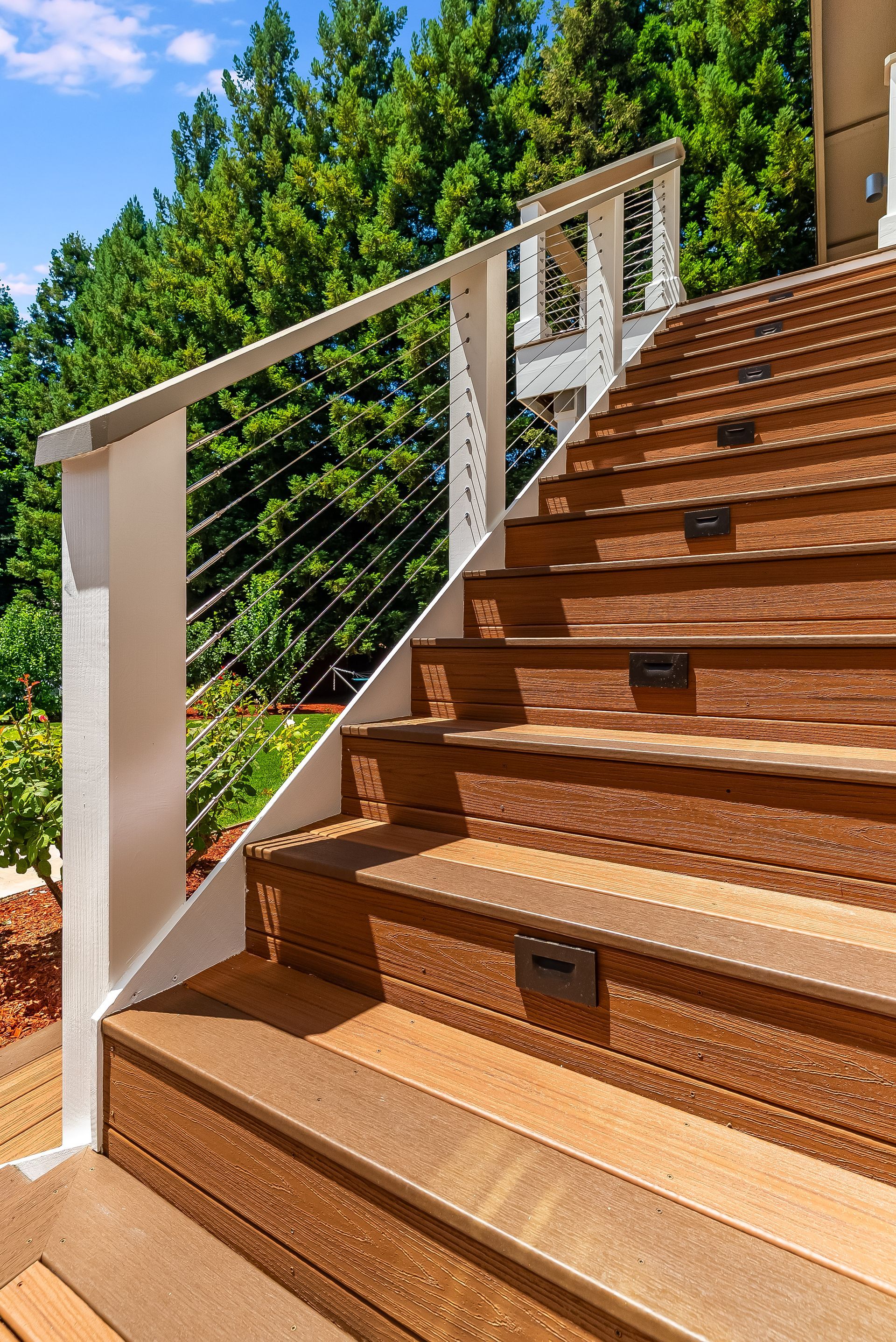Wooden outdoor stairs with white railing beside green trees in bright sunlight
