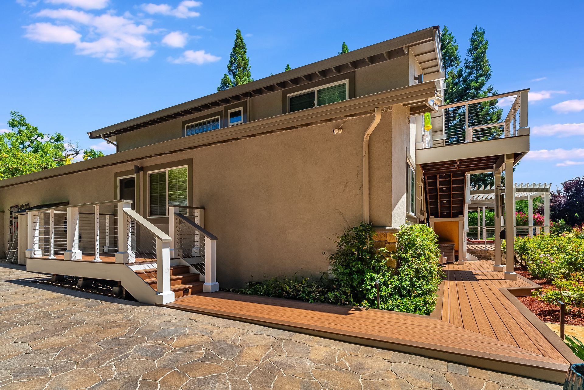 Brown two-story house with a wooden deck, white railings, and a brick patio in sunlight