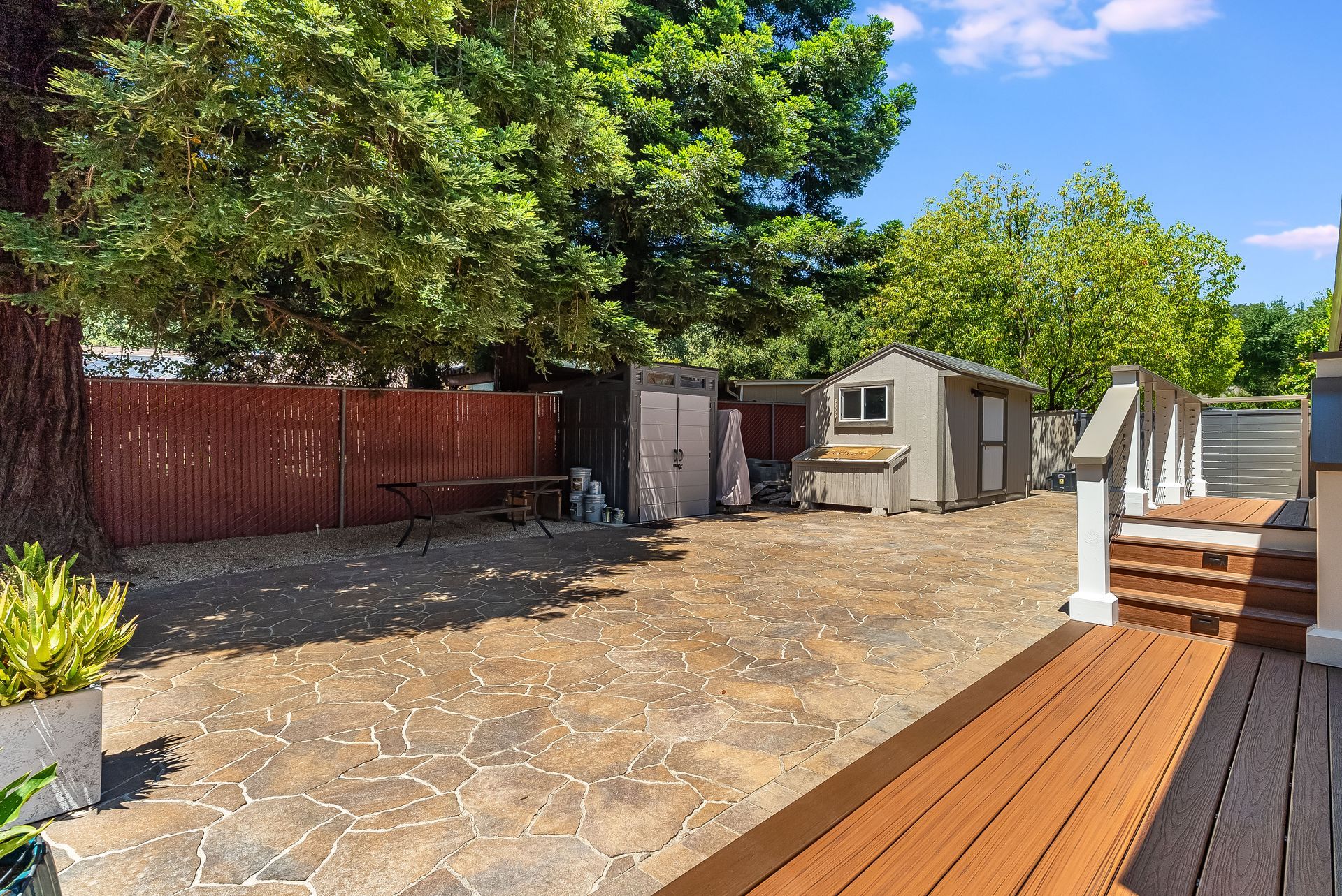 Backyard patio with stone paving, wooden deck, garden shed, and trees under a blue sky