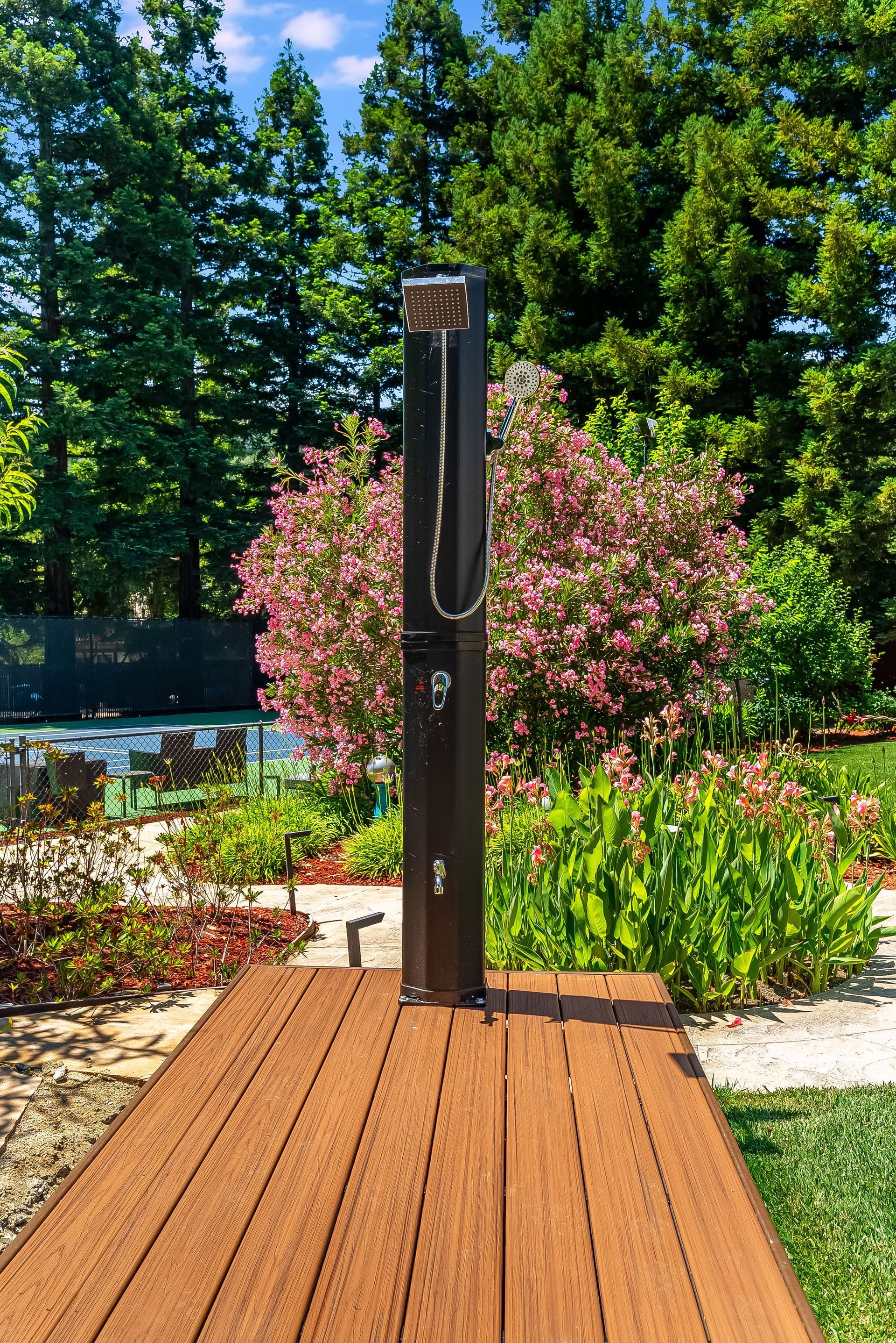Outdoor shower on a wooden deck in a landscaped garden with trees and pink flowers.