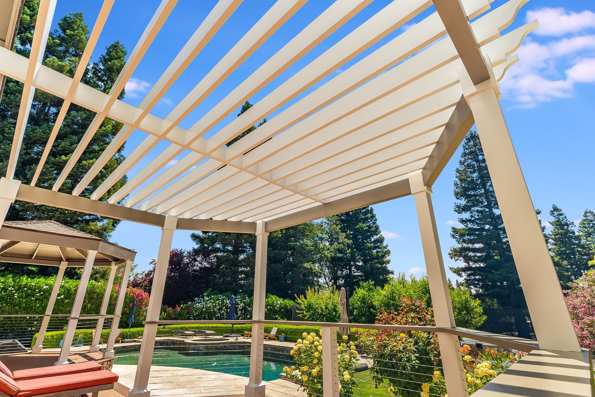 Pergola frame over backyard patio with pool, trees, and bright blue sky