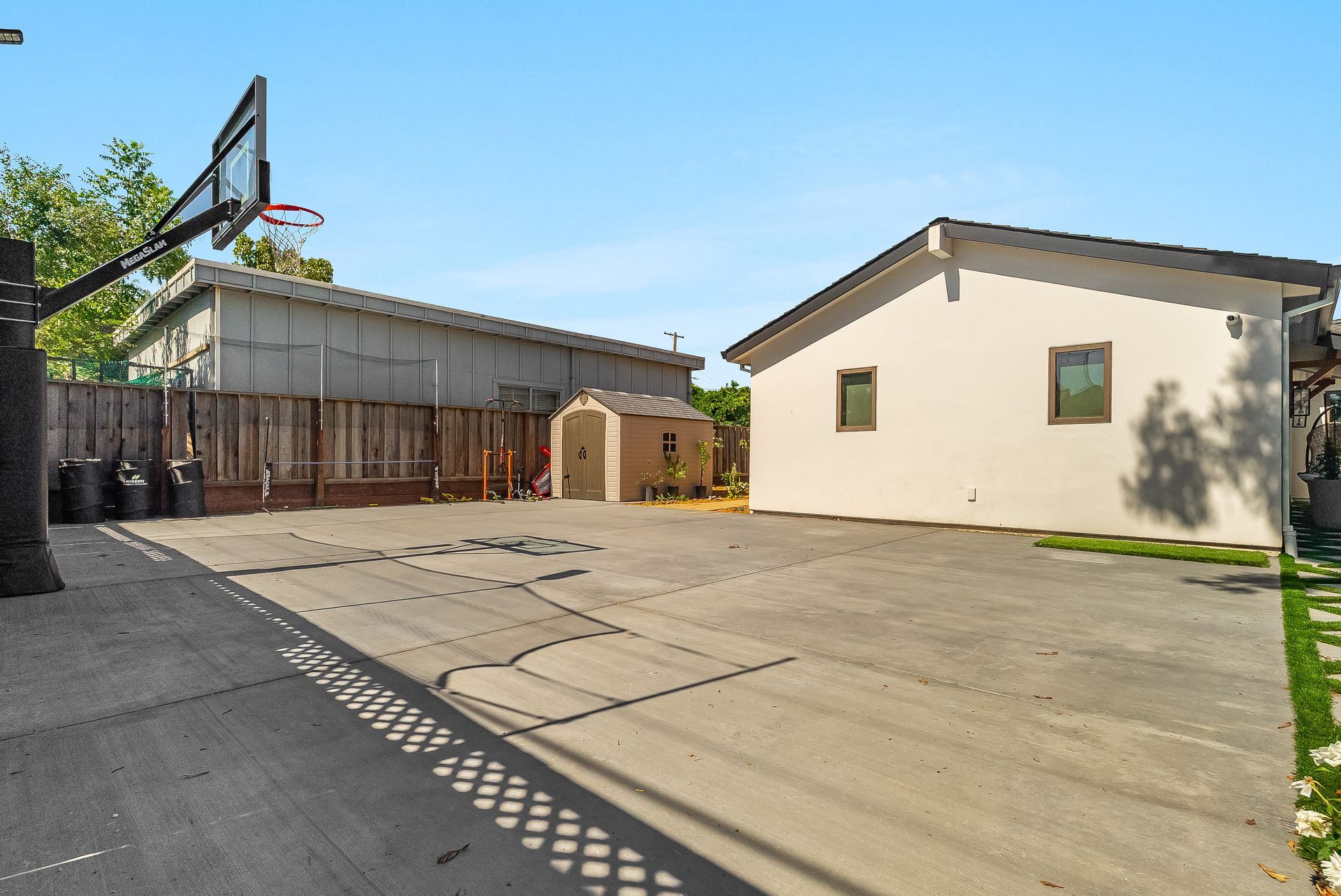 Backyard with basketball hoop, concrete driveway, wooden fence, and beige garage in sunlight