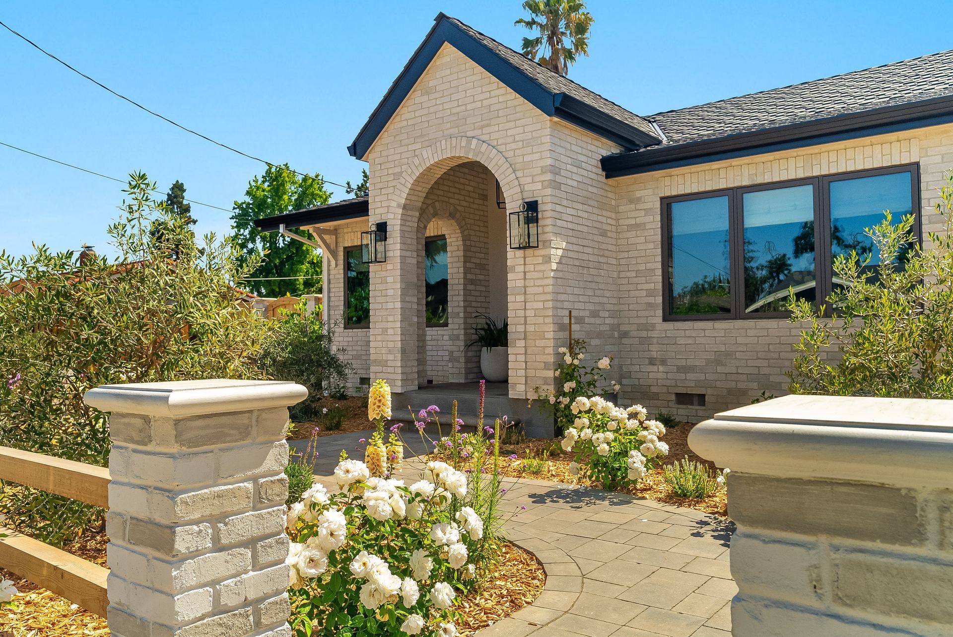 Single-story tan brick house with arched entry, blue windows, and a landscaped front garden under a clear sky