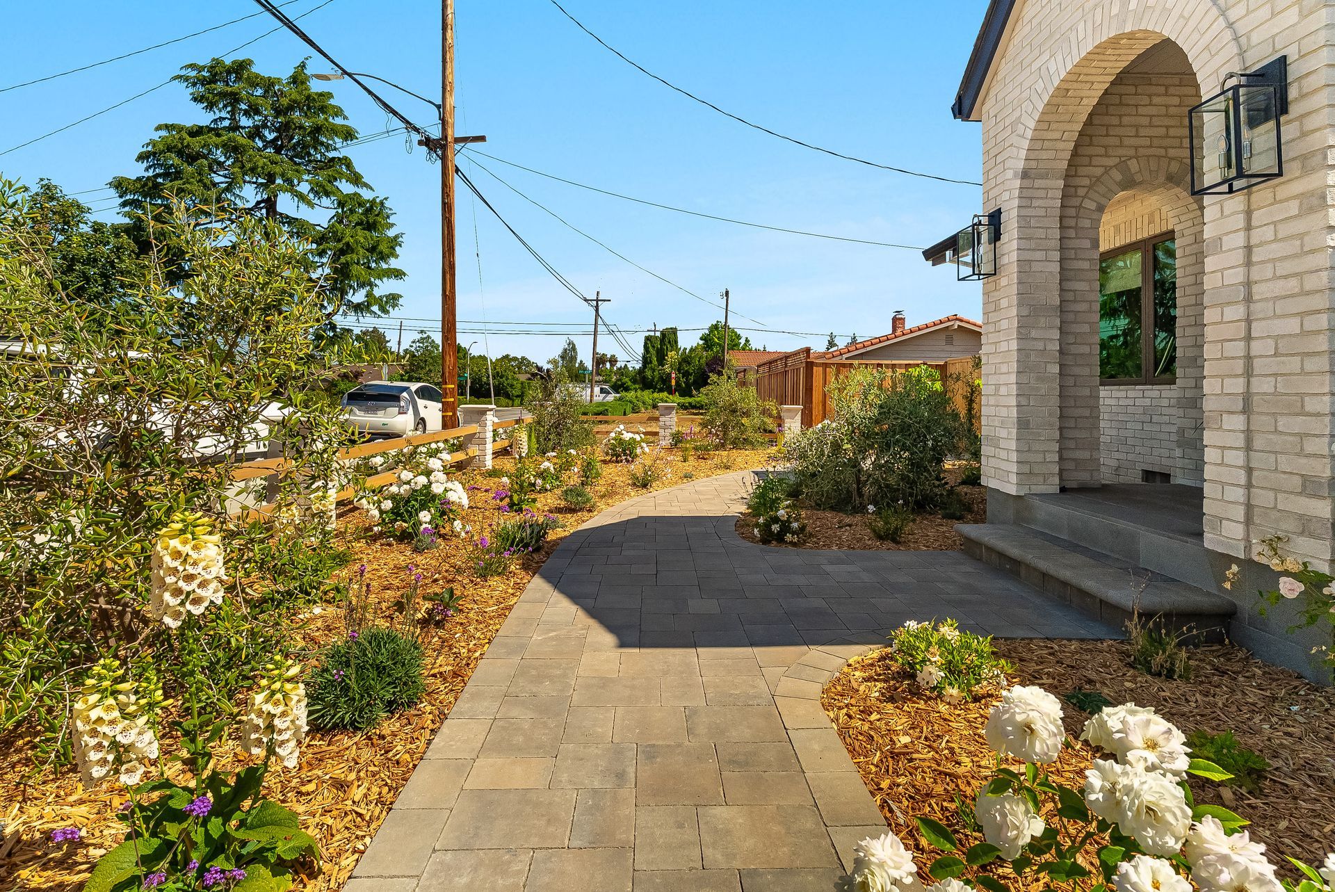 Sunlit garden walkway beside a stone house, with flowers, shrubs, and a utility pole under a blue sky