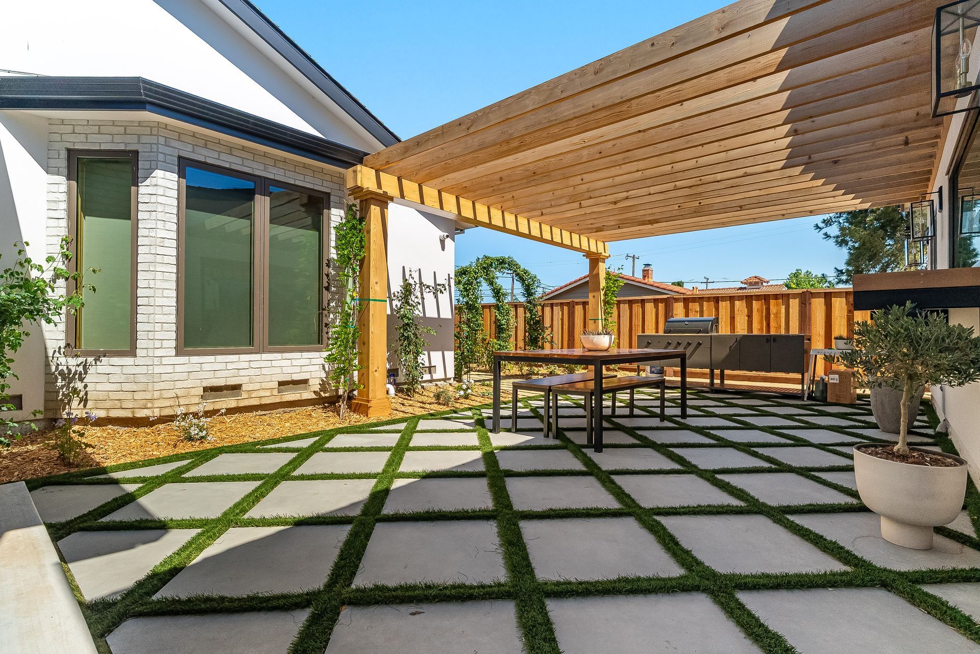 Modern patio with stone pavers, pergola shade, outdoor dining table, and potted plants beside a house