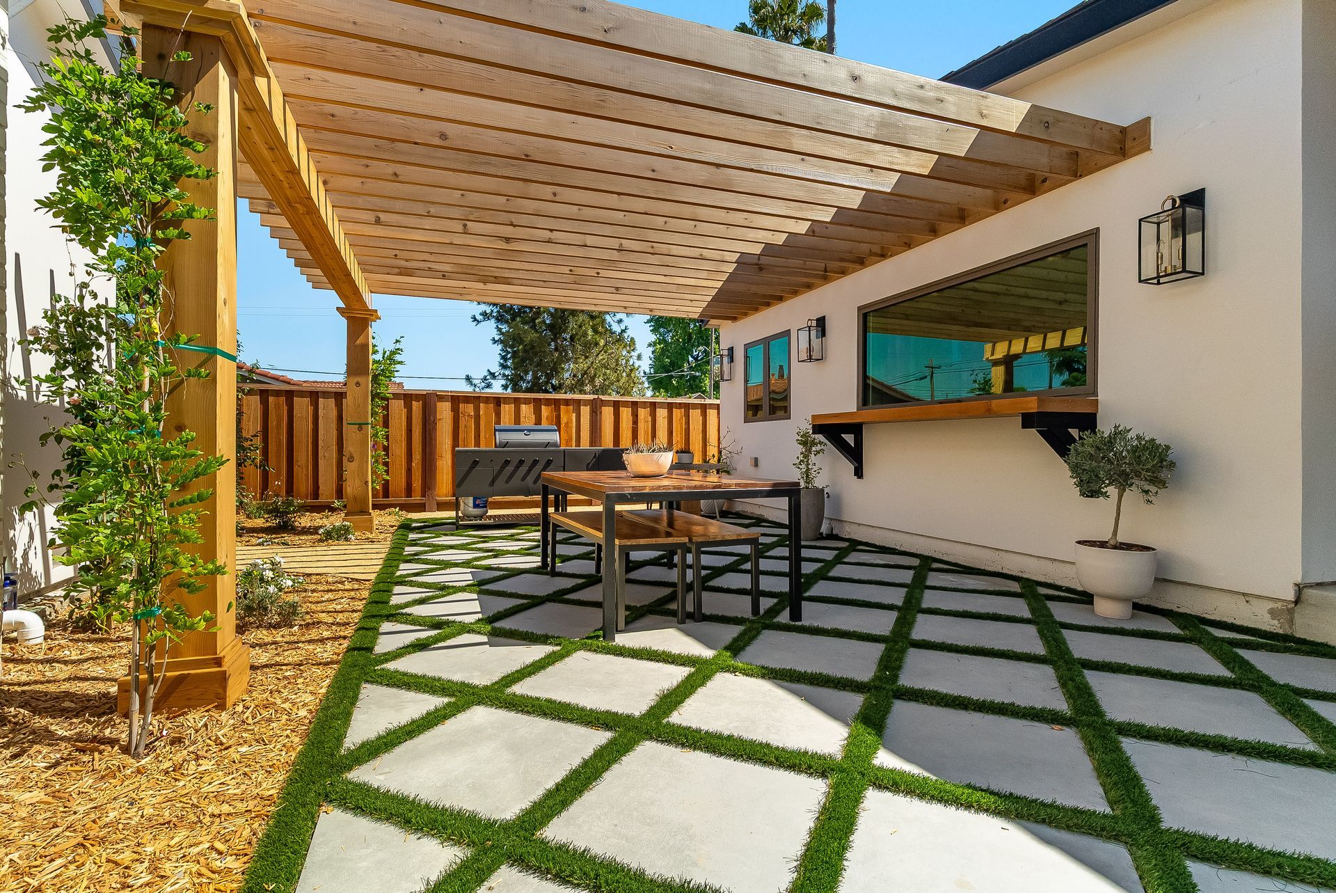 Modern patio with pergola, stone pavers, outdoor table, and wooden fence beside a white house