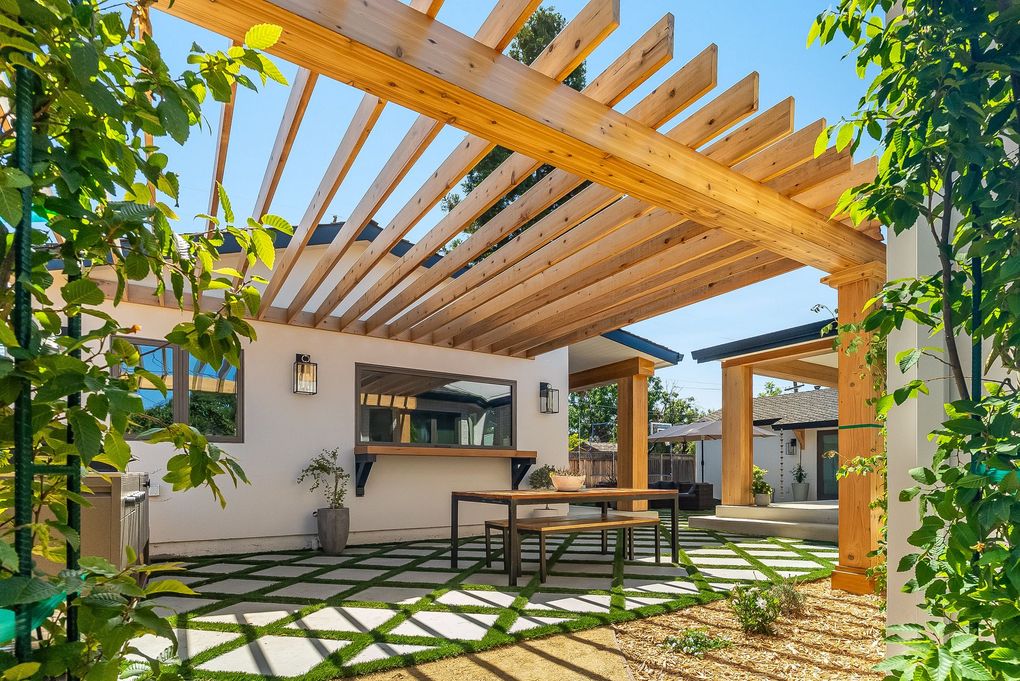 Sunlit patio with wooden pergola, dining table, and lush greenery beside a modern home