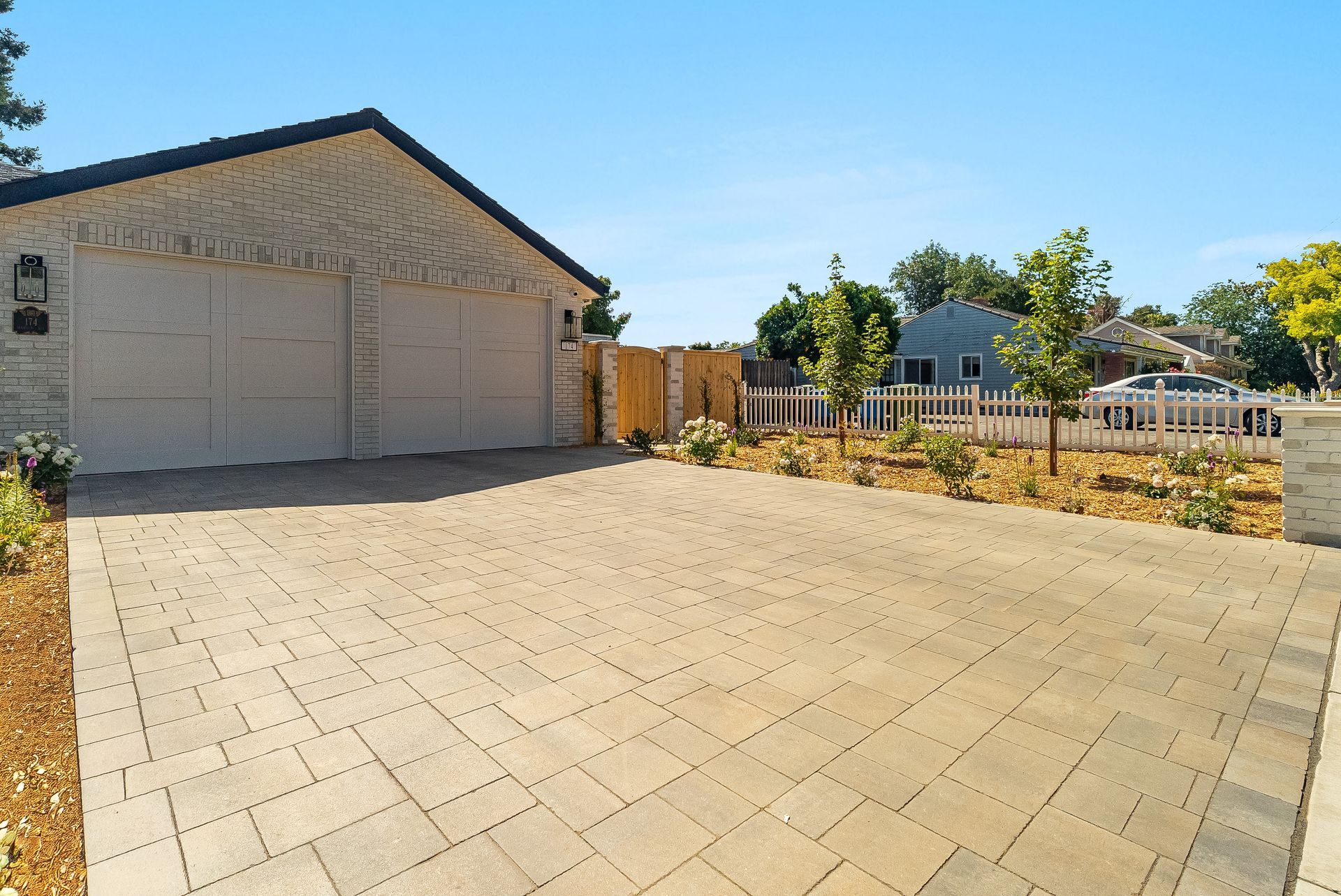 Sunny paved driveway beside a garage and fenced backyard with trees and a small house