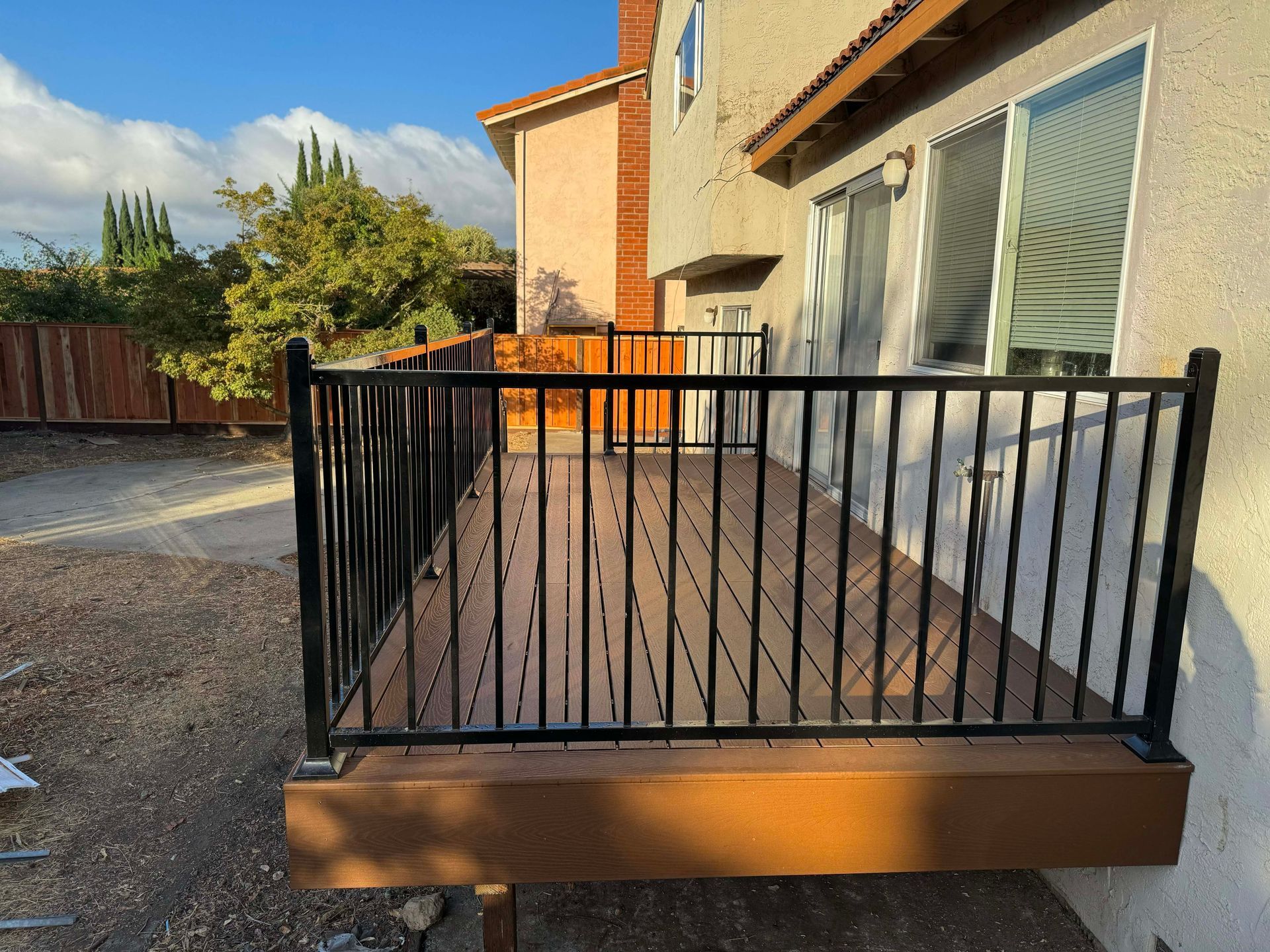 A brown raised wooden deck with a black metal railing attached to the side of a stucco house.