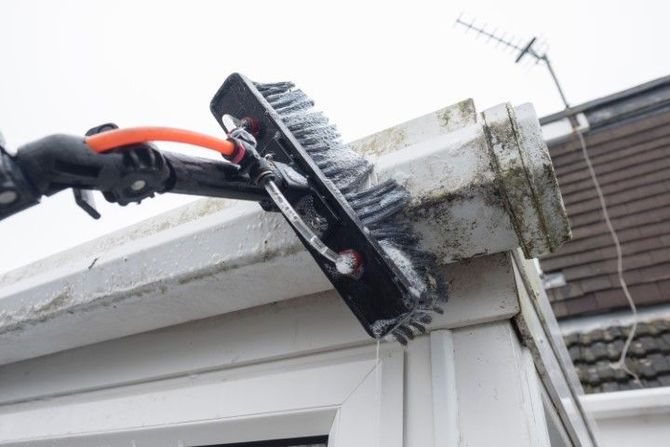 Gloved hand using a brush to clean the roof edge and gutter on a house