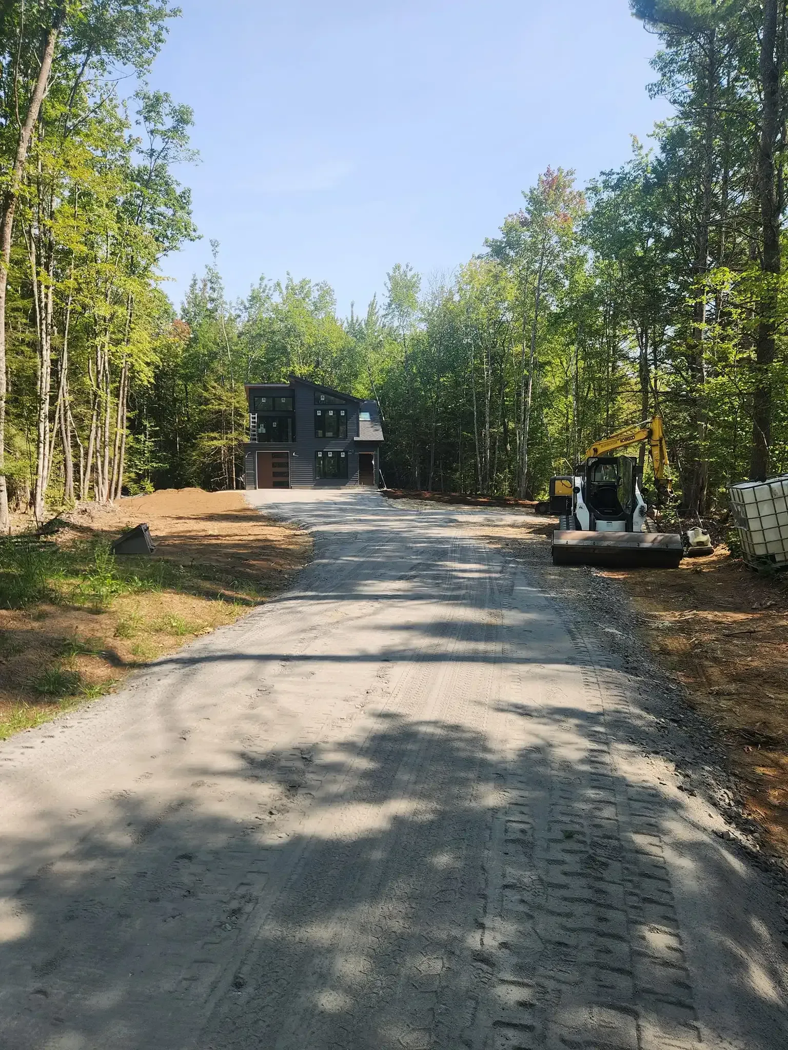 Gravel driveway leads to a modern black house nestled in a forest. Construction equipment sits on the right.