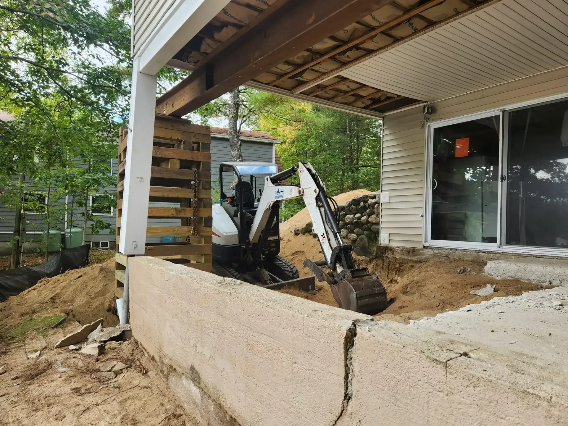 Mini excavator digging near a house foundation. Construction site with dirt, wooden supports, and sliding glass door.