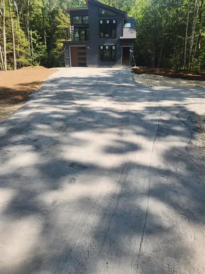 Modern gray house with gravel driveway, surrounded by trees.