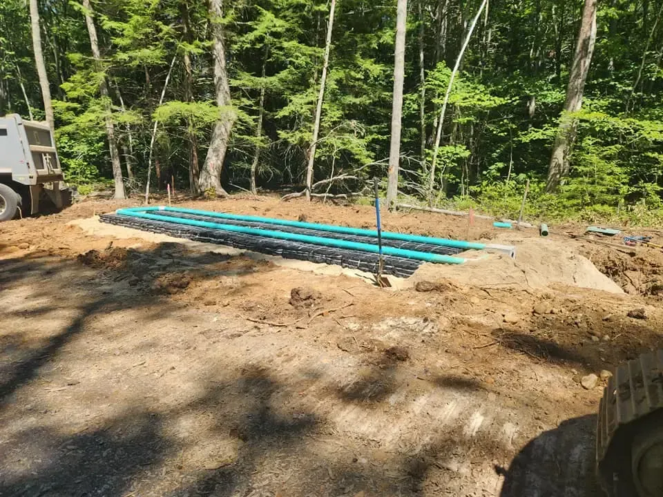 Construction site with exposed blue piping in a rectangular trench, surrounded by dirt and trees.
