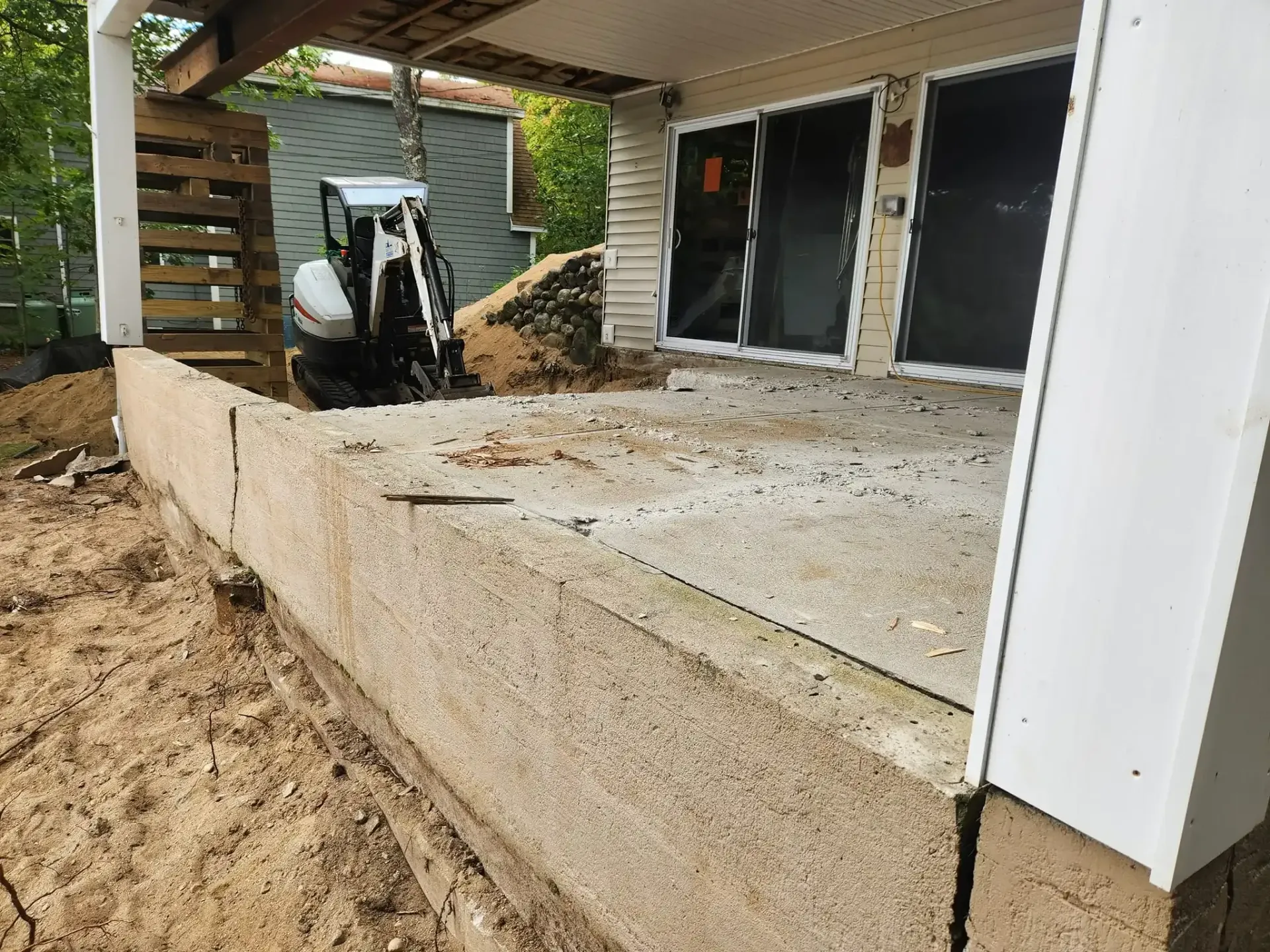 Construction site with mini-excavator digging near a porch with sliding glass doors; exterior shot.
