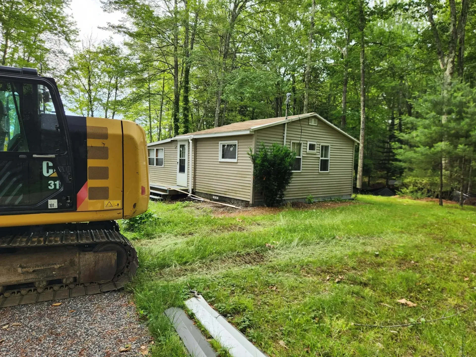 Yellow excavator next to a small, single-story house in a wooded area. House has gray siding, green grass.