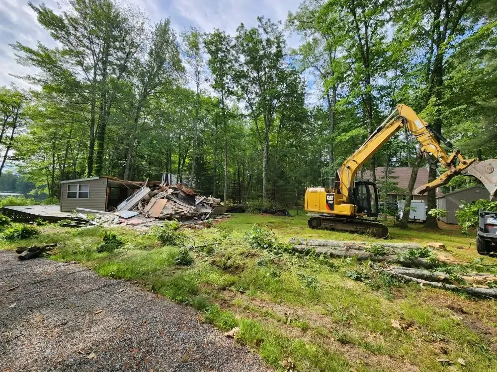 Yellow excavator demolishing a building; debris on the ground; trees in the background.