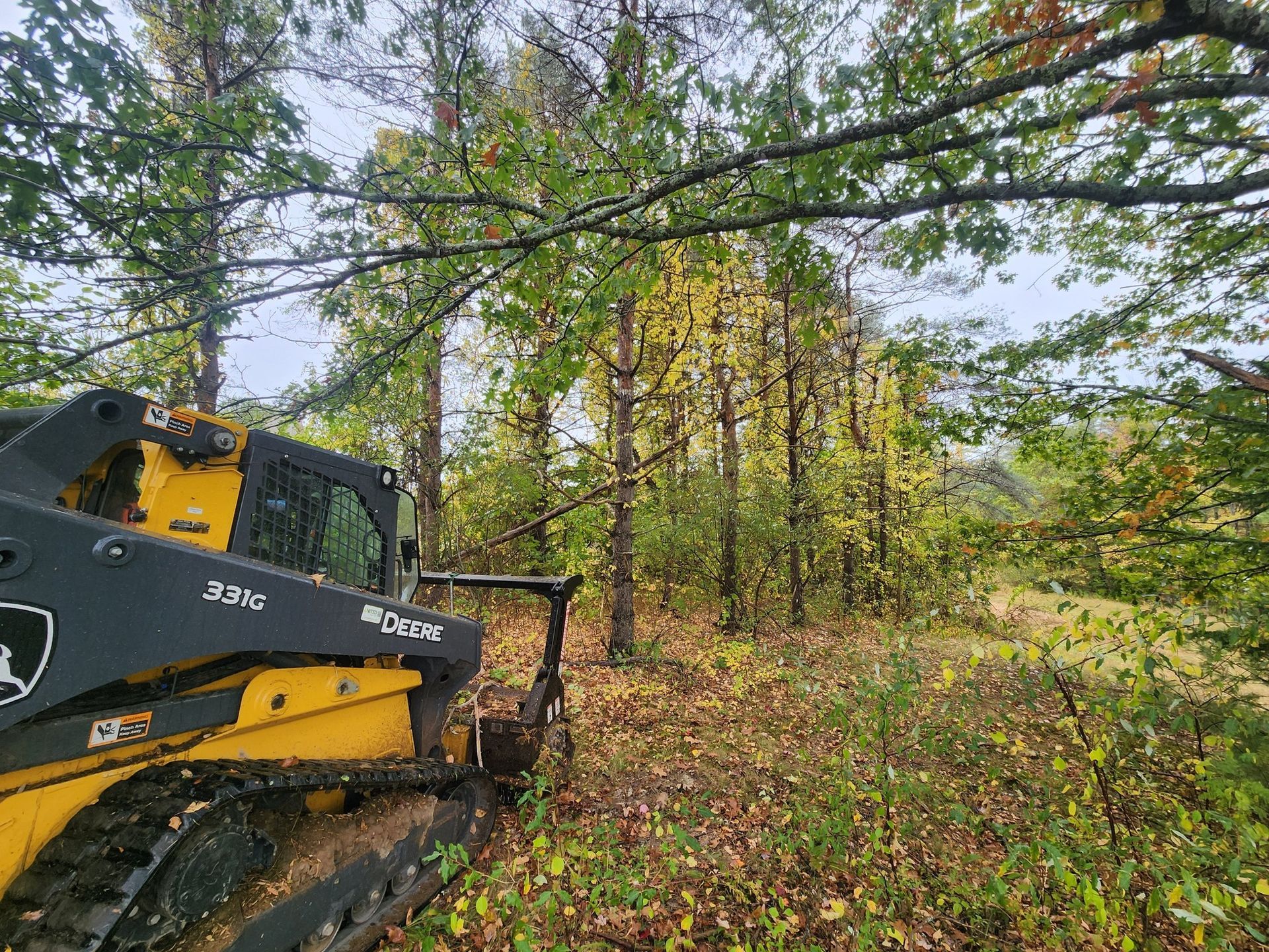 Yellow John Deere skid steer clearing trees in wooded area.