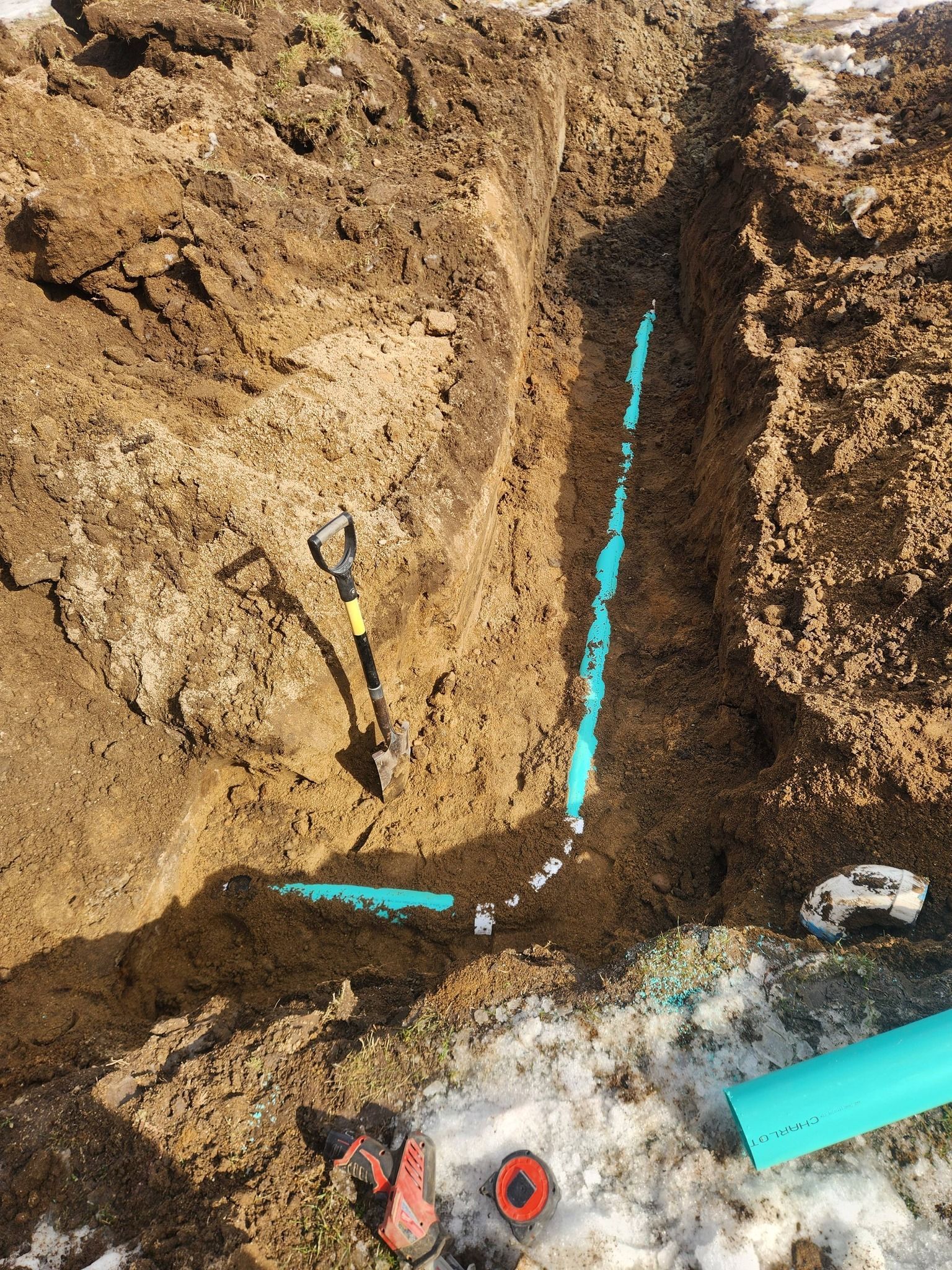 A blue PVC pipe is laid inside a narrow trench in the earth, featuring an angled joint. Tools lie nearby in the snow.