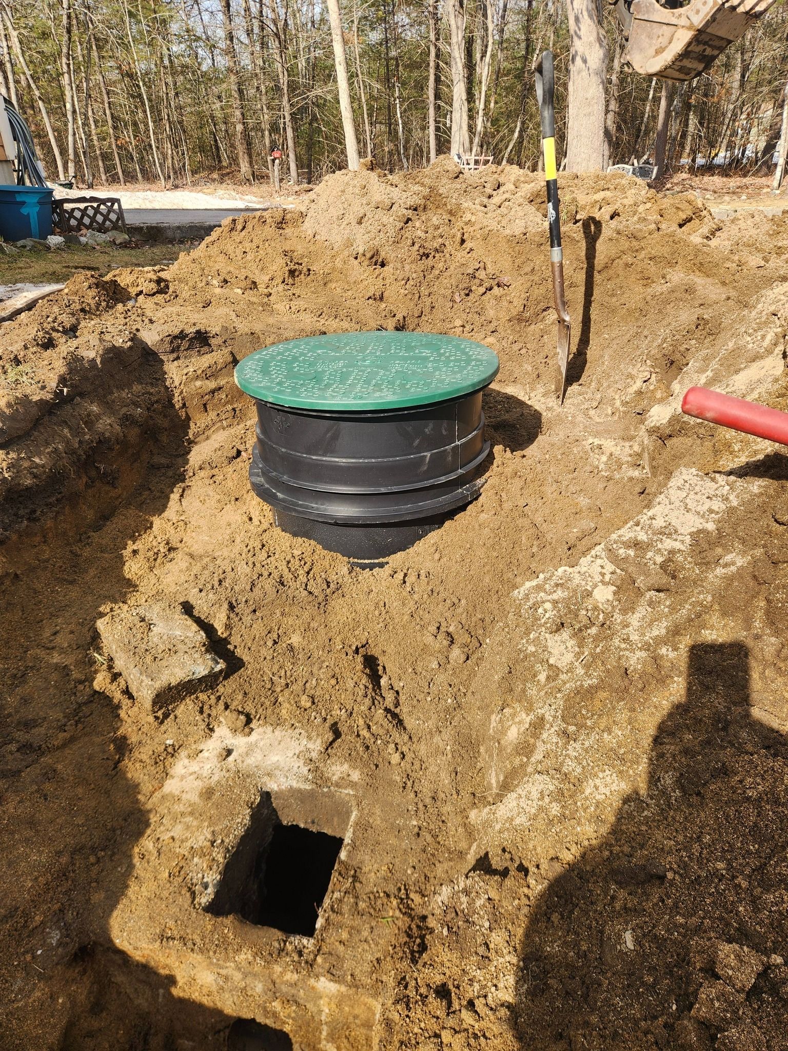 A green septic tank riser lid sits in an excavated dirt pit with an exposed concrete opening nearby.