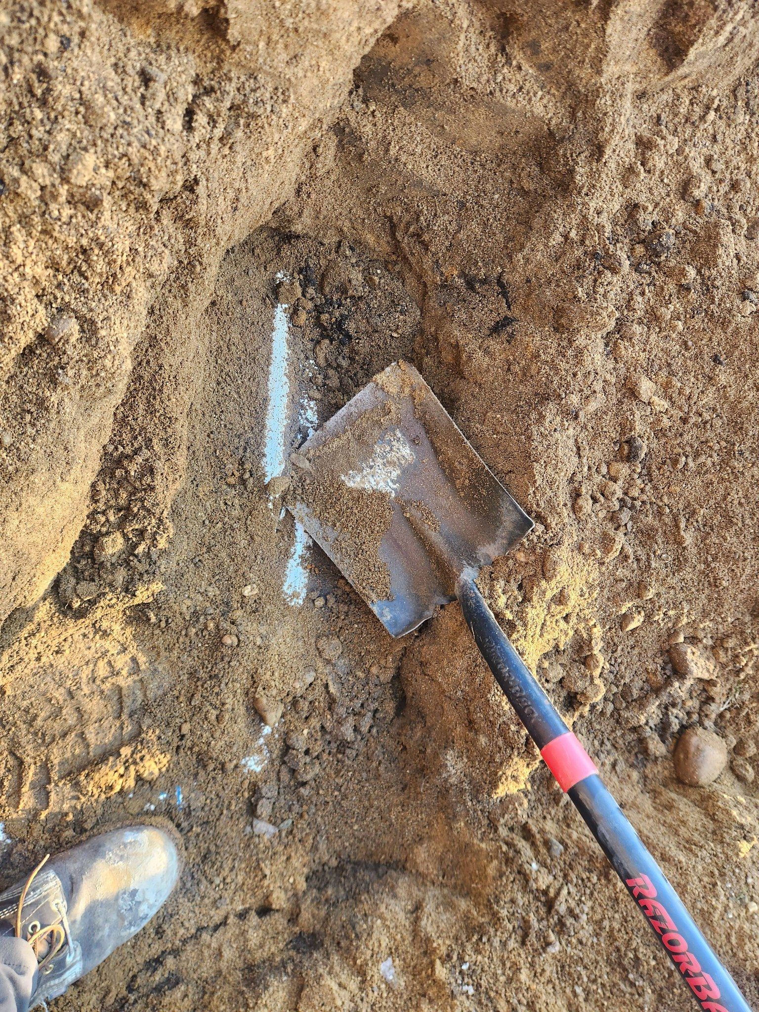 A shovel rests in a dirt hole next to a white utility pipe exposed in the ground.