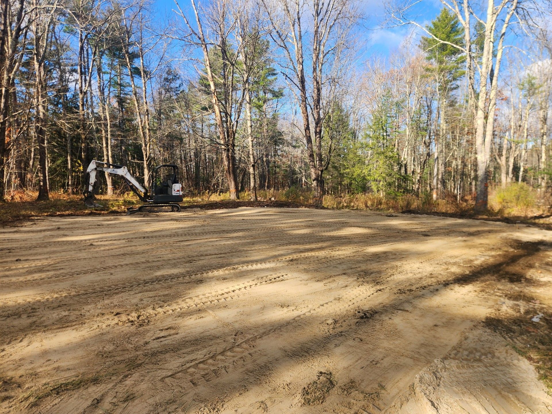 Cleared sandy area with excavator, trees in the background under a blue sky.