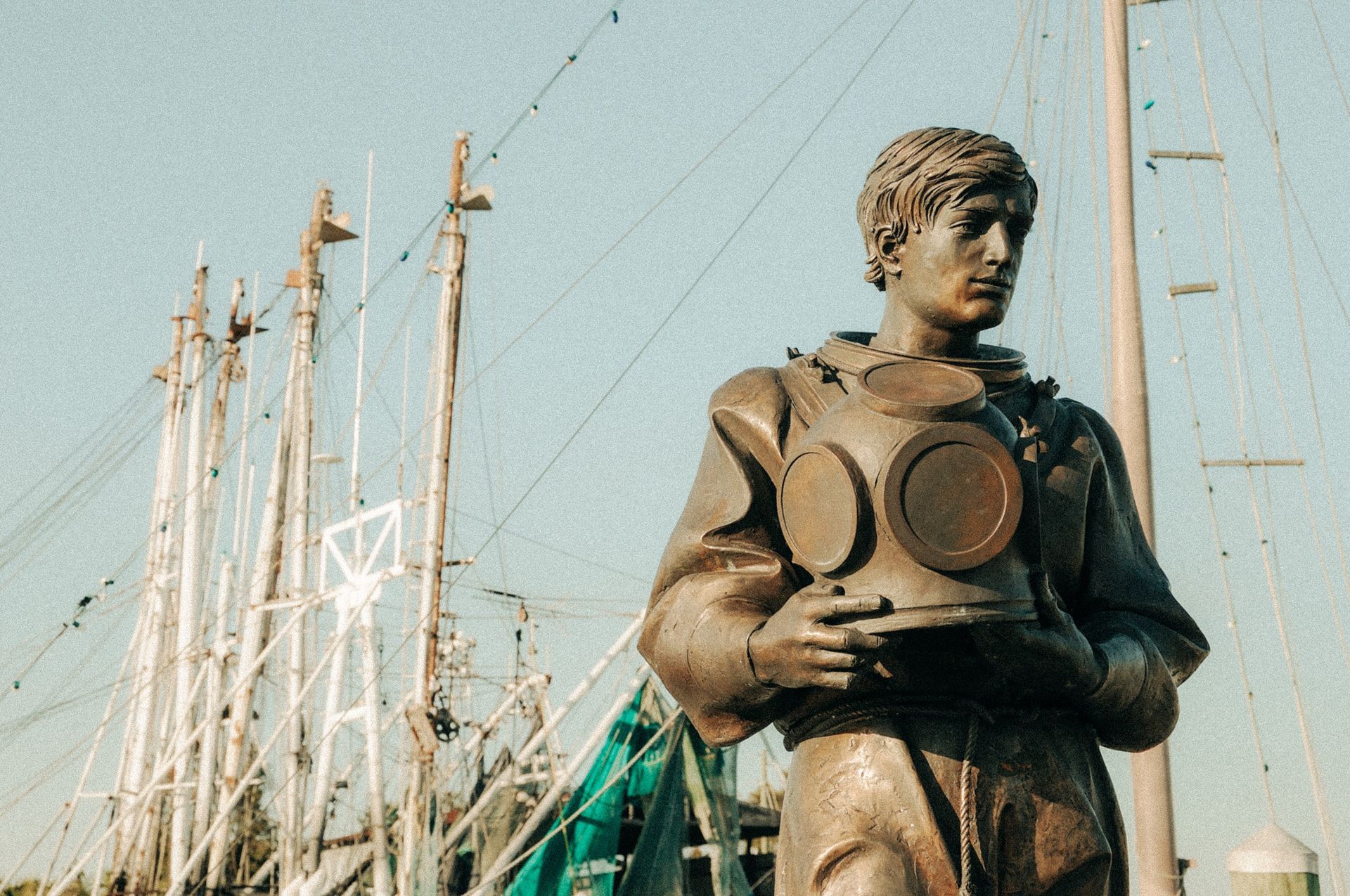 Bronze diver statue holding a helmet near sailboat masts under a pale blue sky