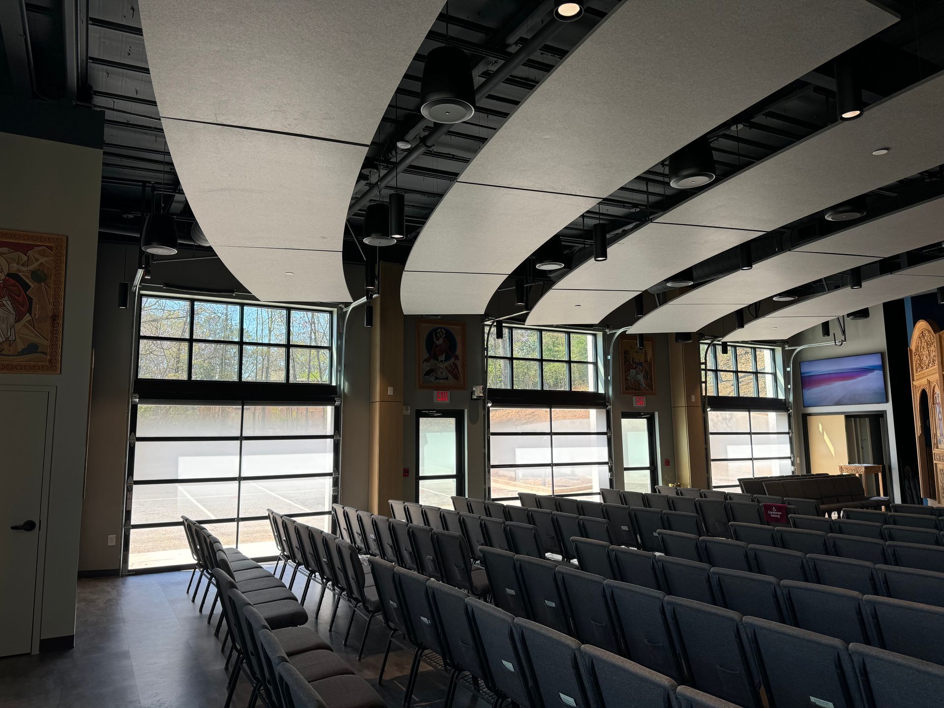 Empty auditorium with rows of gray seats, curved white ceiling panels, and large windows at the back.