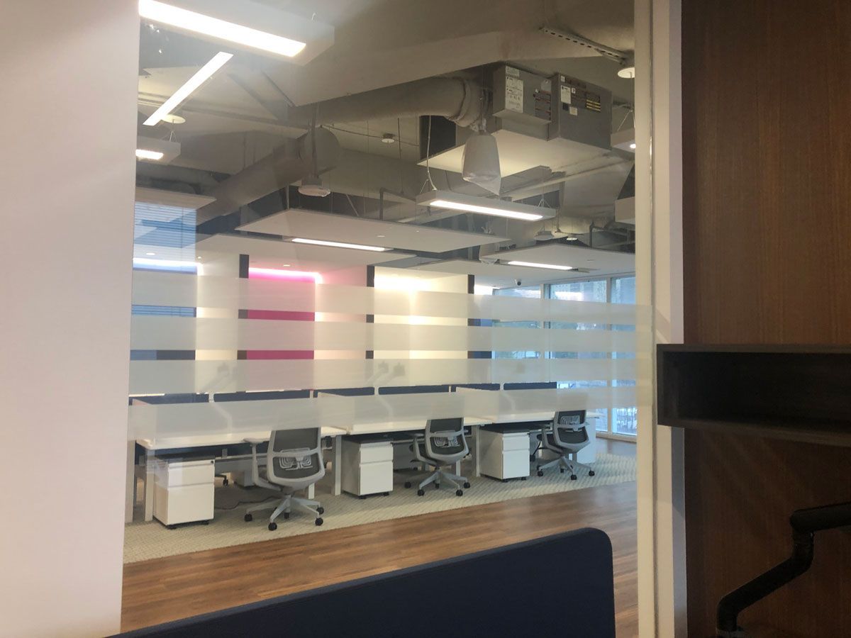Modern office meeting room seen through glass, with chairs, desks, and ceiling lights.