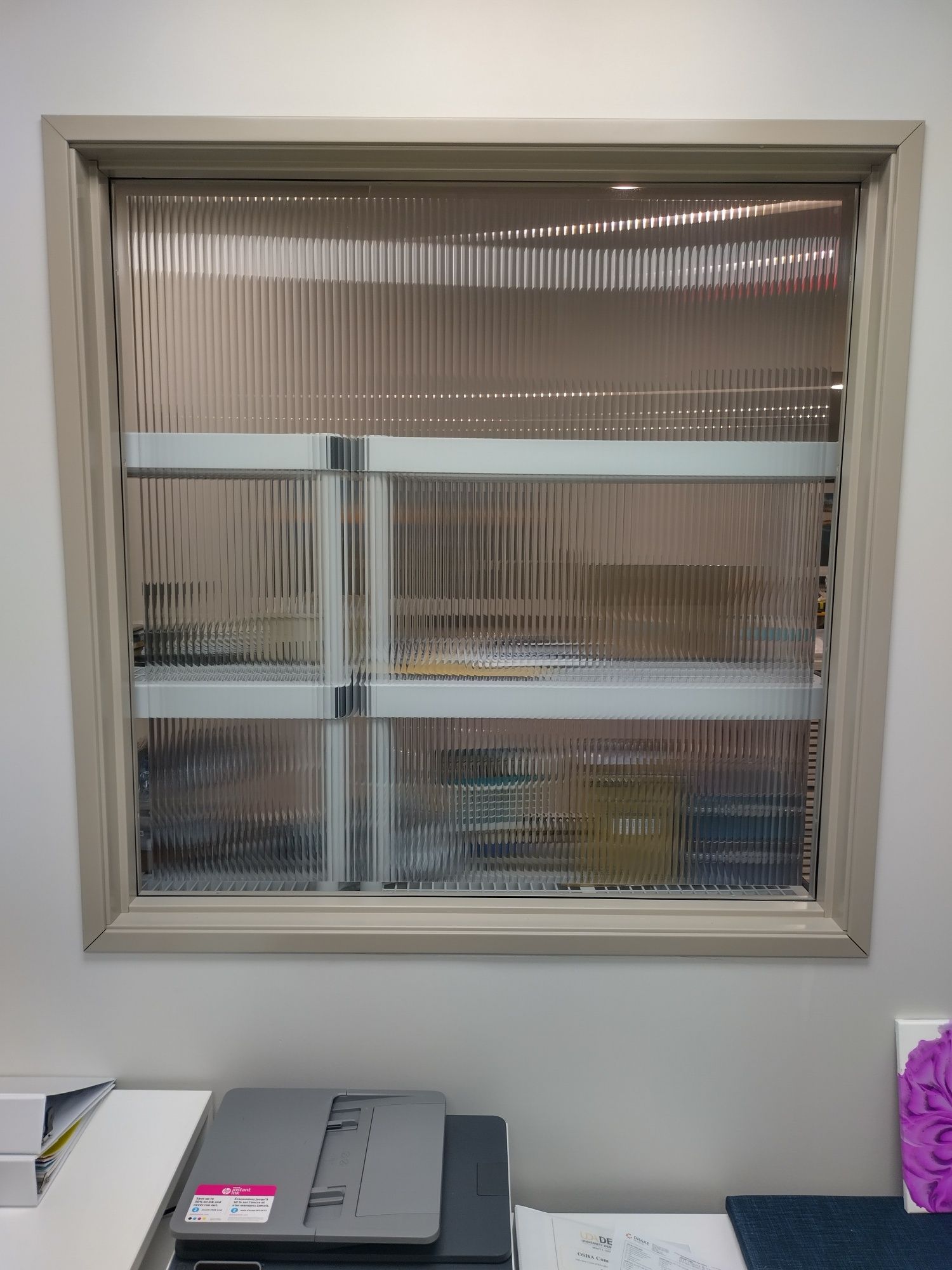 Small office window with textured glass above a printer on a desk, and a purple item at right.