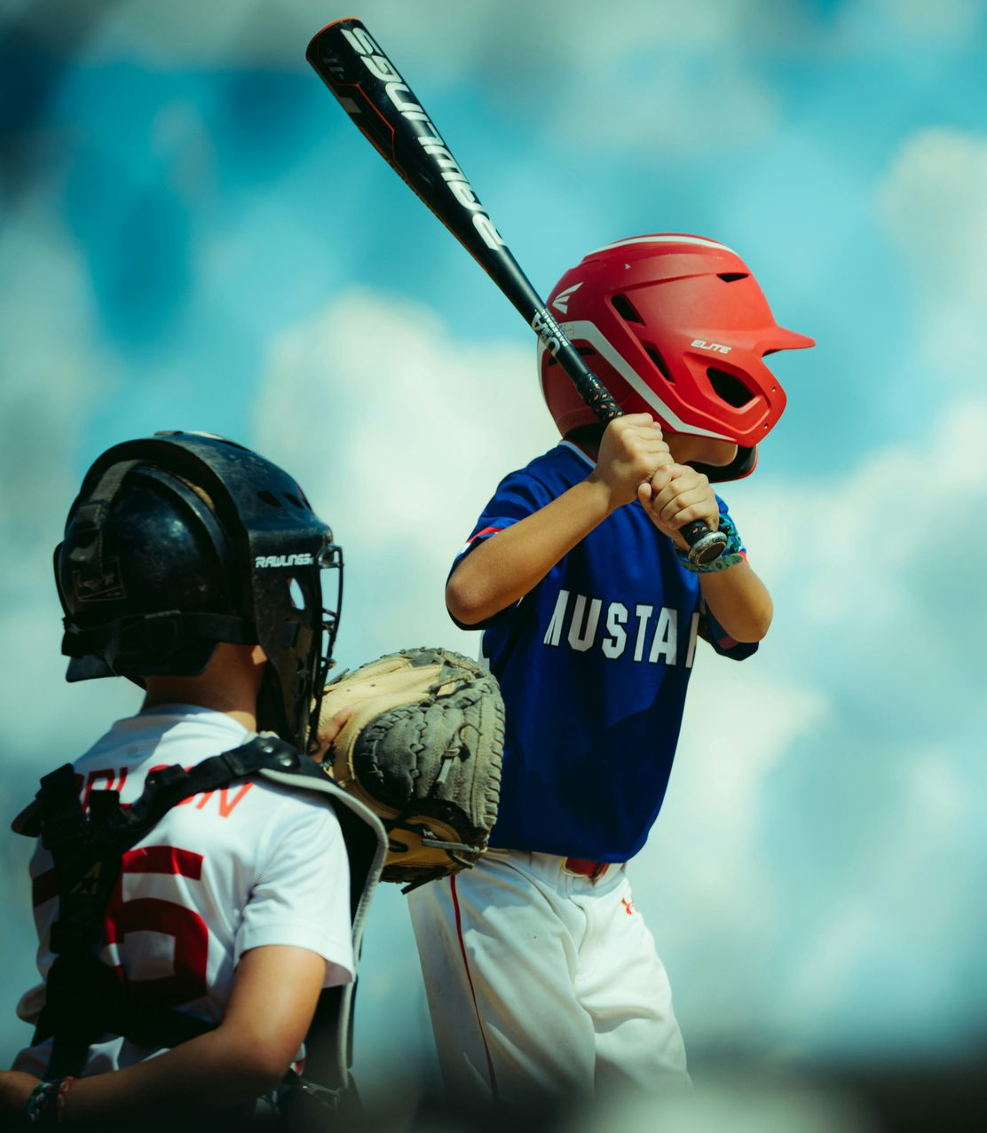 A young baseball batter in a blue uniform and red helmet stands at home plate, with a catcher positioned behind.