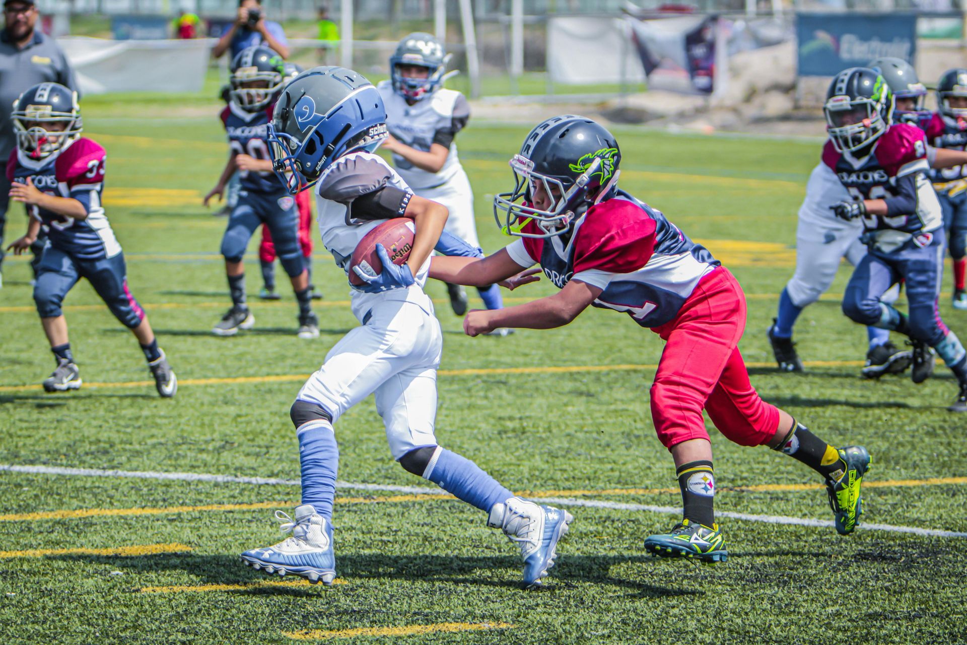 Youth football players in uniforms compete on a green field as one player in white carries the ball while being tackled.